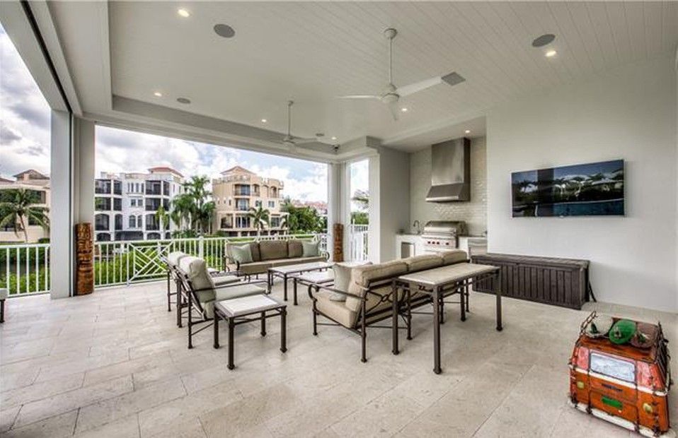 Covered outdoor patio with seating, grilling area, TV, overlooking a body of water and buildings.