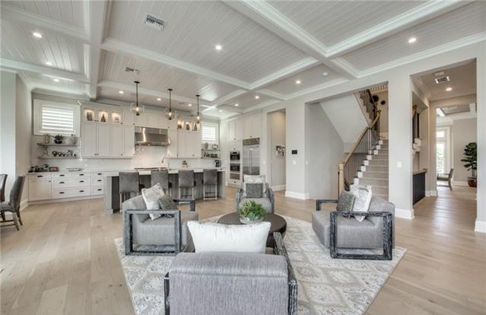 Open-concept living space with kitchen, seating area, and staircase; light wood floors, gray furniture, white cabinets.