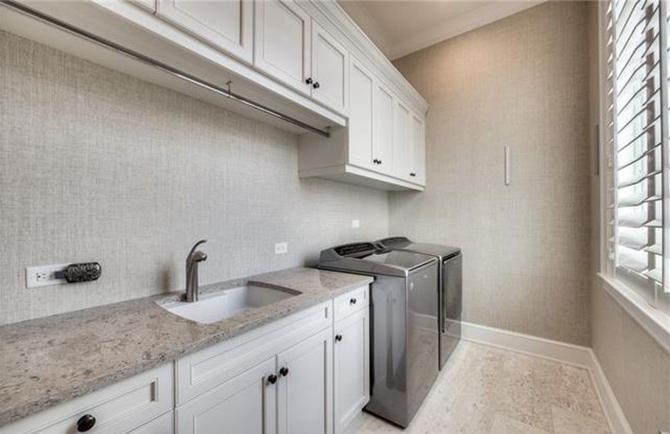 Laundry room with white cabinets, stainless steel washer/dryer, sink, and window with blinds.
