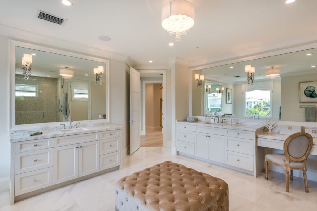 Spacious, bright white bathroom with large mirrors, marble countertops, and an upholstered ottoman.