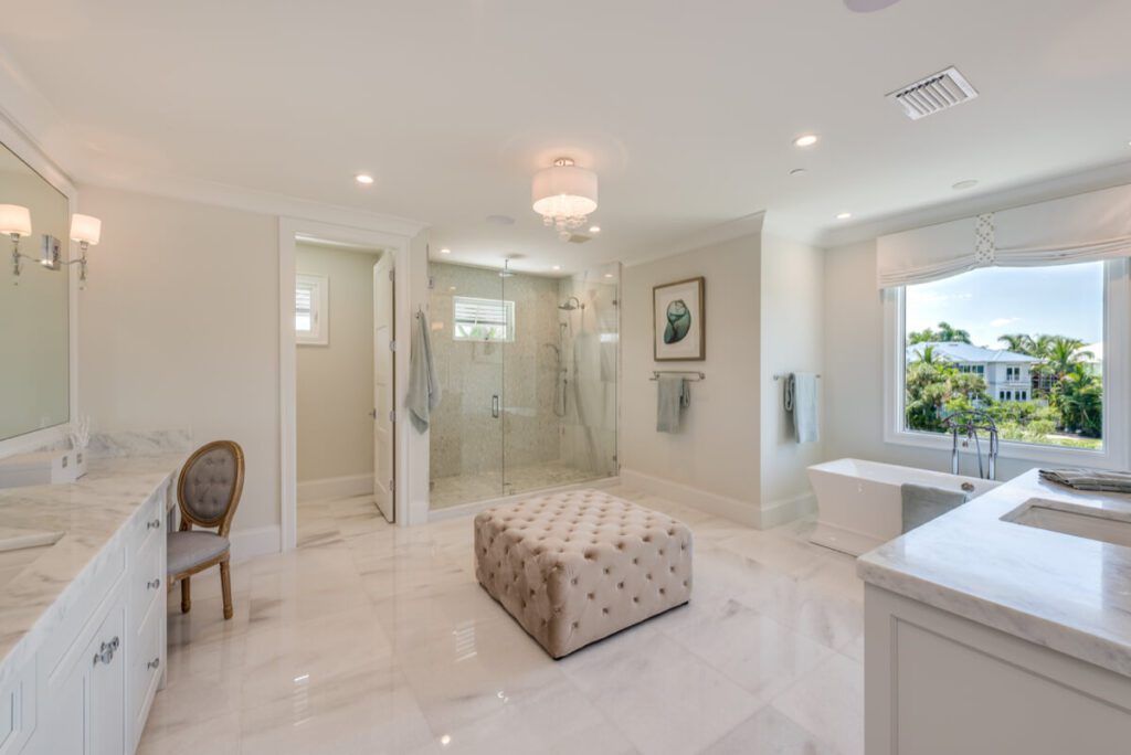 Spacious, bright white bathroom with marble floors, walk-in shower, soaking tub, vanity, and large ottoman.