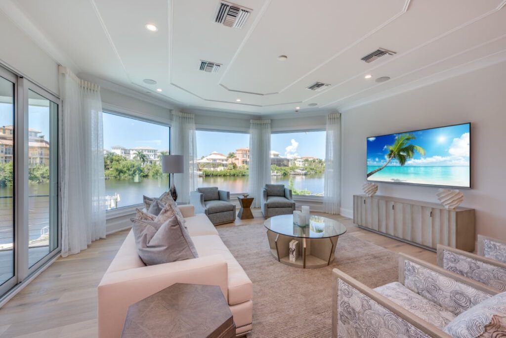 Spacious living room with waterfront view, white walls, large windows, light furniture, and a TV showing a beach scene.