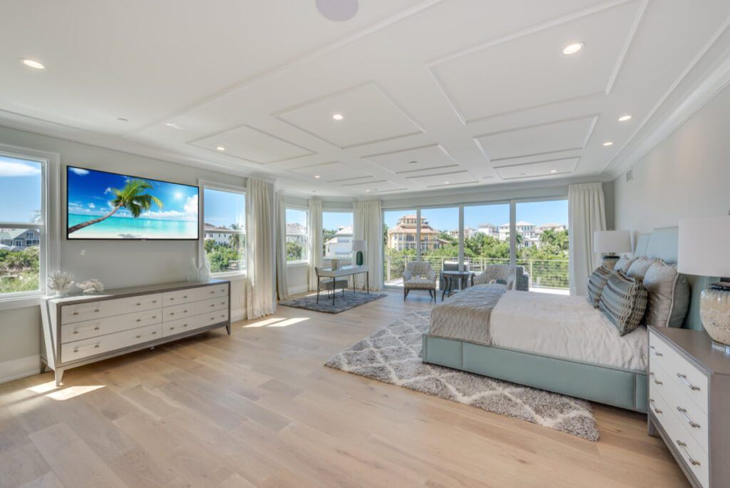 Spacious bedroom with beach view, large windows, TV, light wood floors, and bed with blue accents.