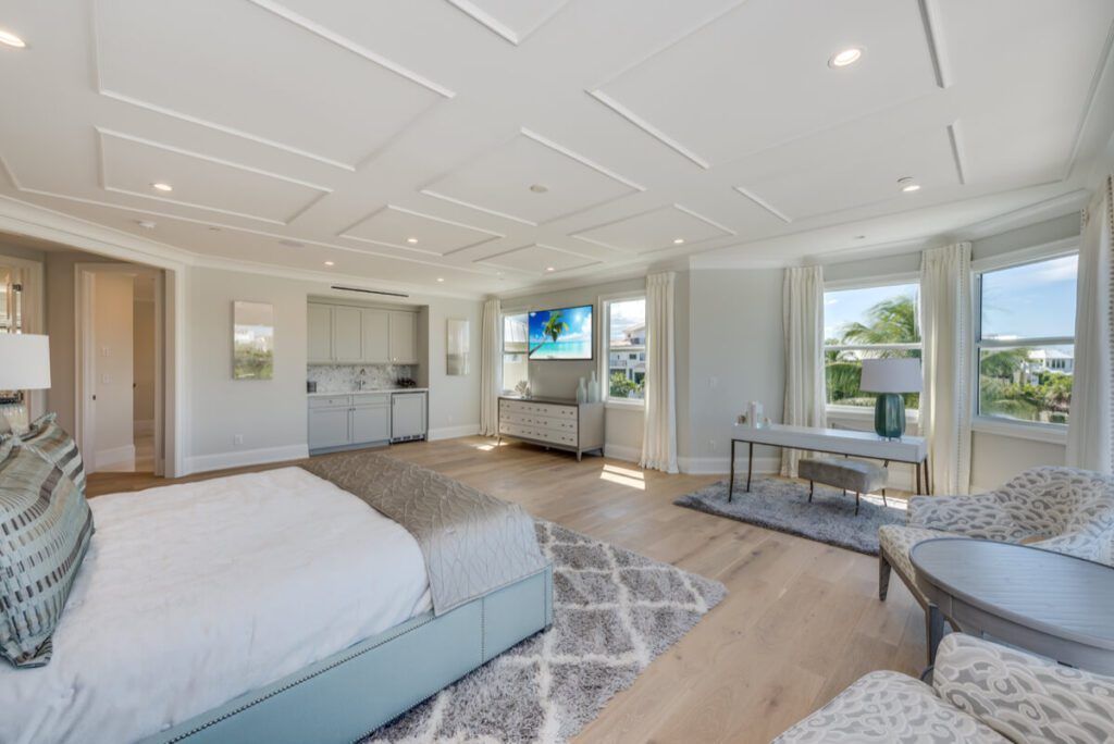 Spacious bedroom with a bed, TV, seating area, and mini-kitchen. Light wood floors, white walls, and a coffered ceiling.