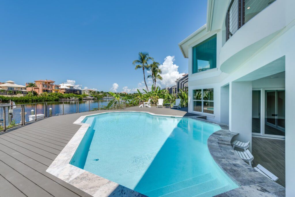 Poolside view of modern white house with blue pool, water, and palm trees under a sunny sky.