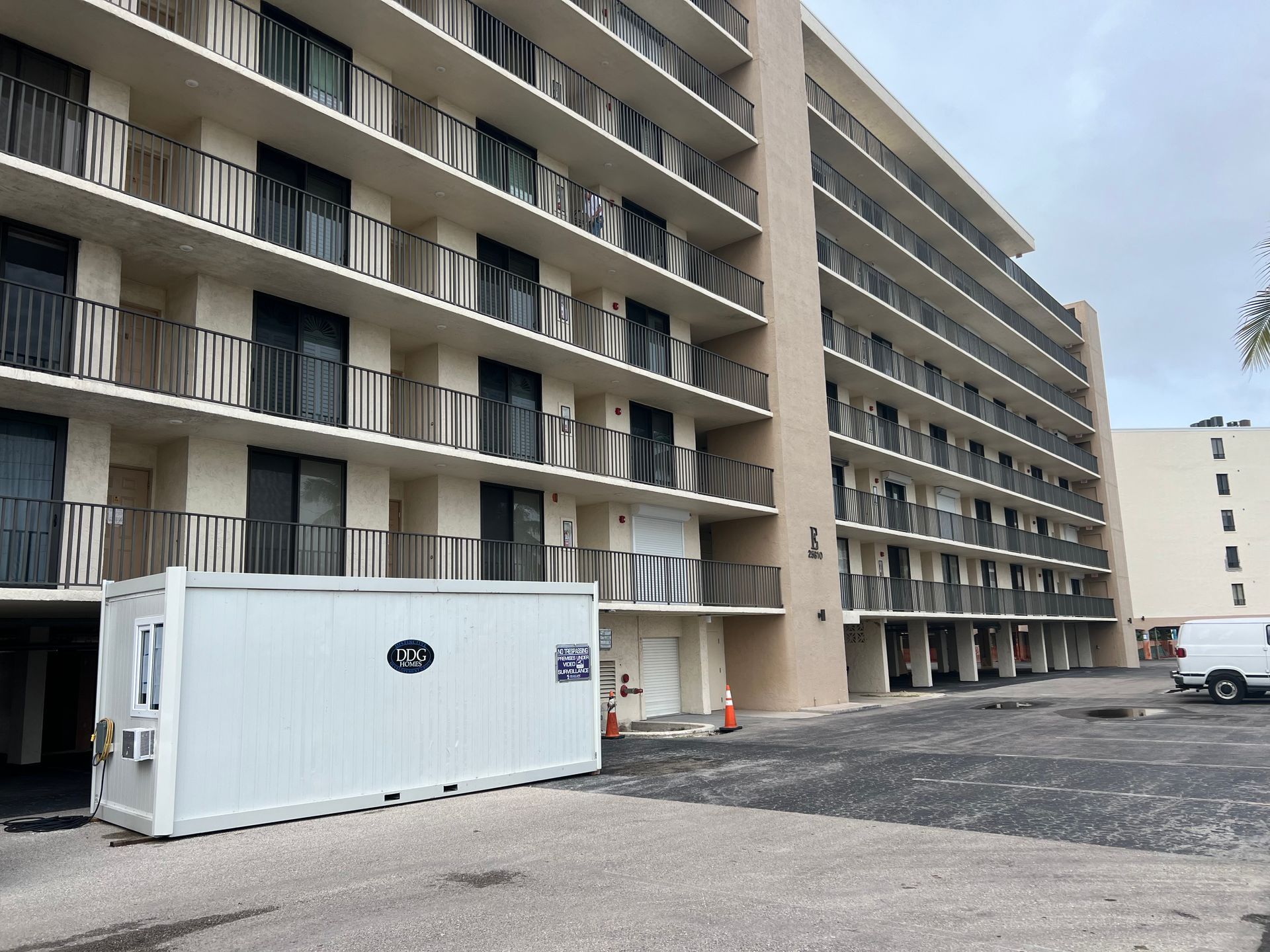 Multi-story beige building with balconies; a white construction trailer sits in the parking lot.