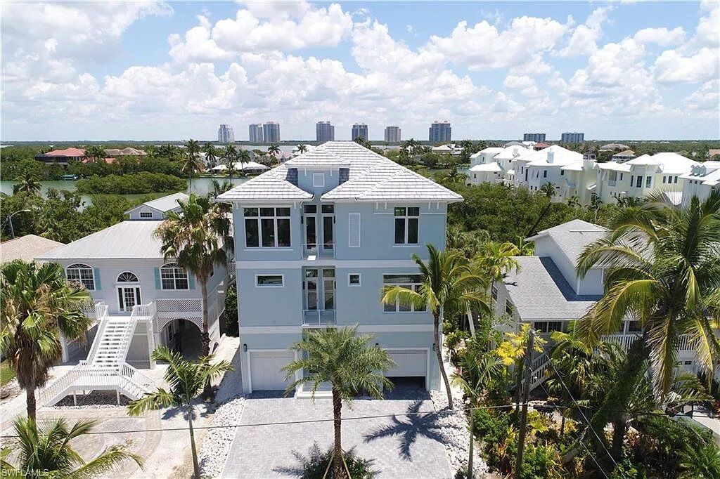 Blue beach house with white roof and garage, palm trees in front, other houses and skyline in the distance.