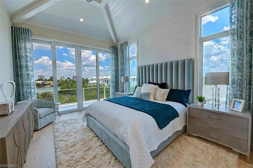 Bedroom with large windows, light blue walls, a bed with a tall headboard, and a fluffy white rug.