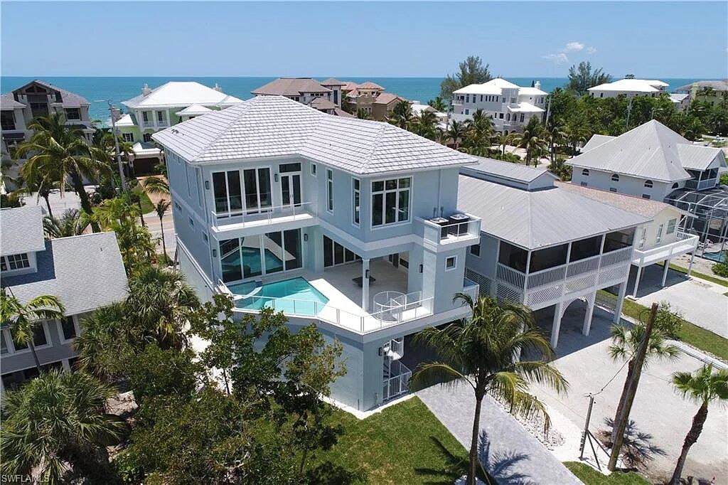 Beachfront light blue house with a pool, ocean view, palm trees, and bright sky.
