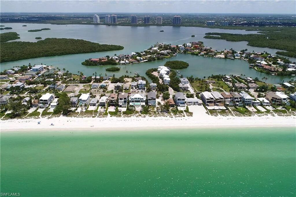 Aerial view of a beach with white sand and turquoise water, houses, and a city skyline in the distance.