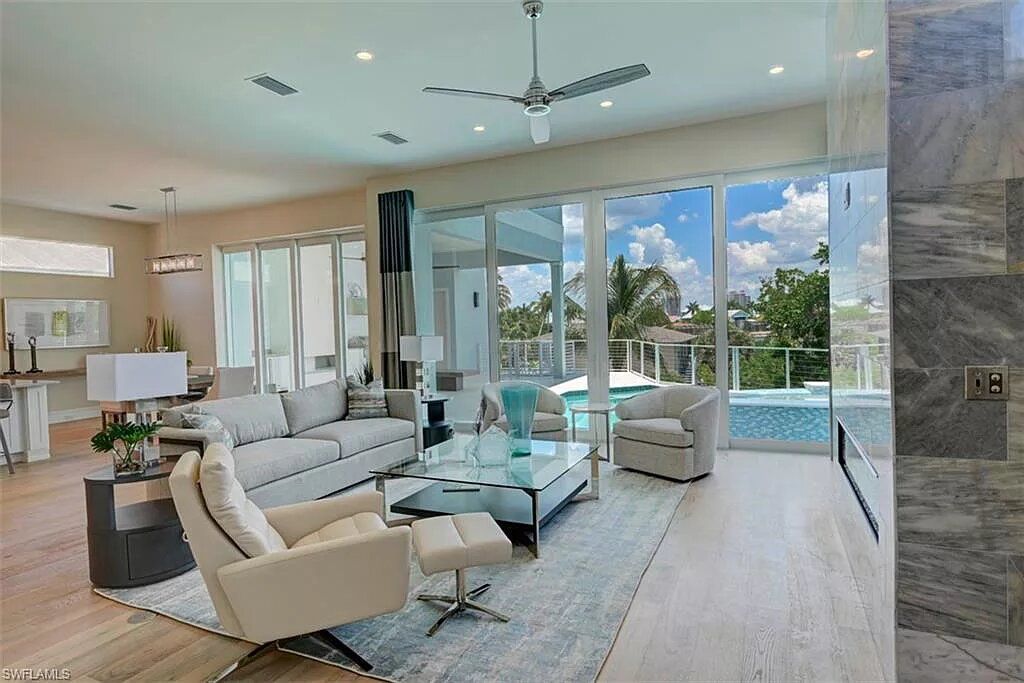 Spacious living room with modern furniture, large windows overlooking a pool, light-colored walls and floors.