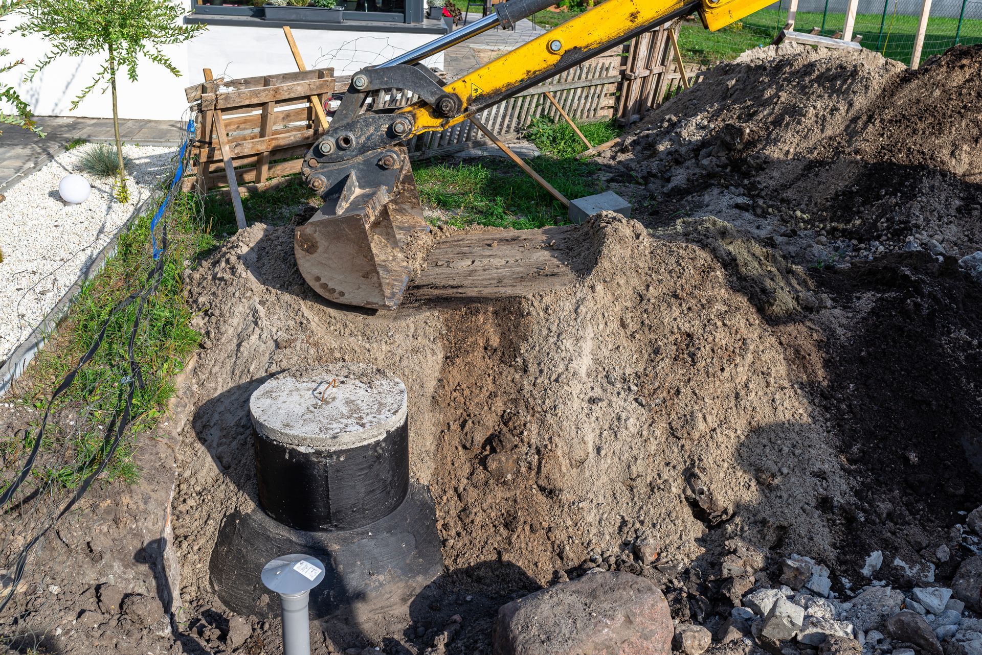 Mini excavator digging near a concrete cylindrical structure, soil pile, and small landscaping.