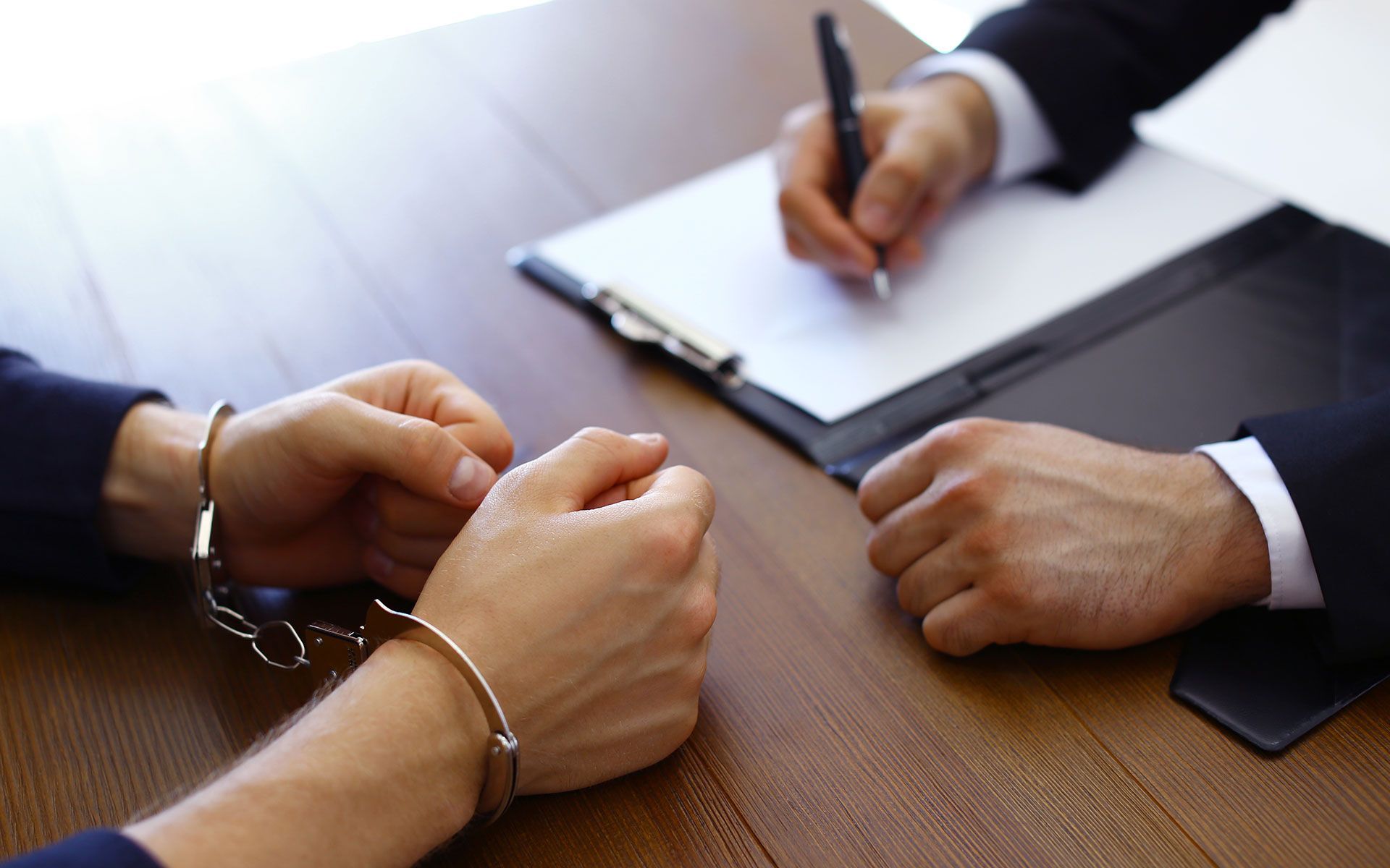 Person in handcuffs at a table, facing a person writing on a clipboard.