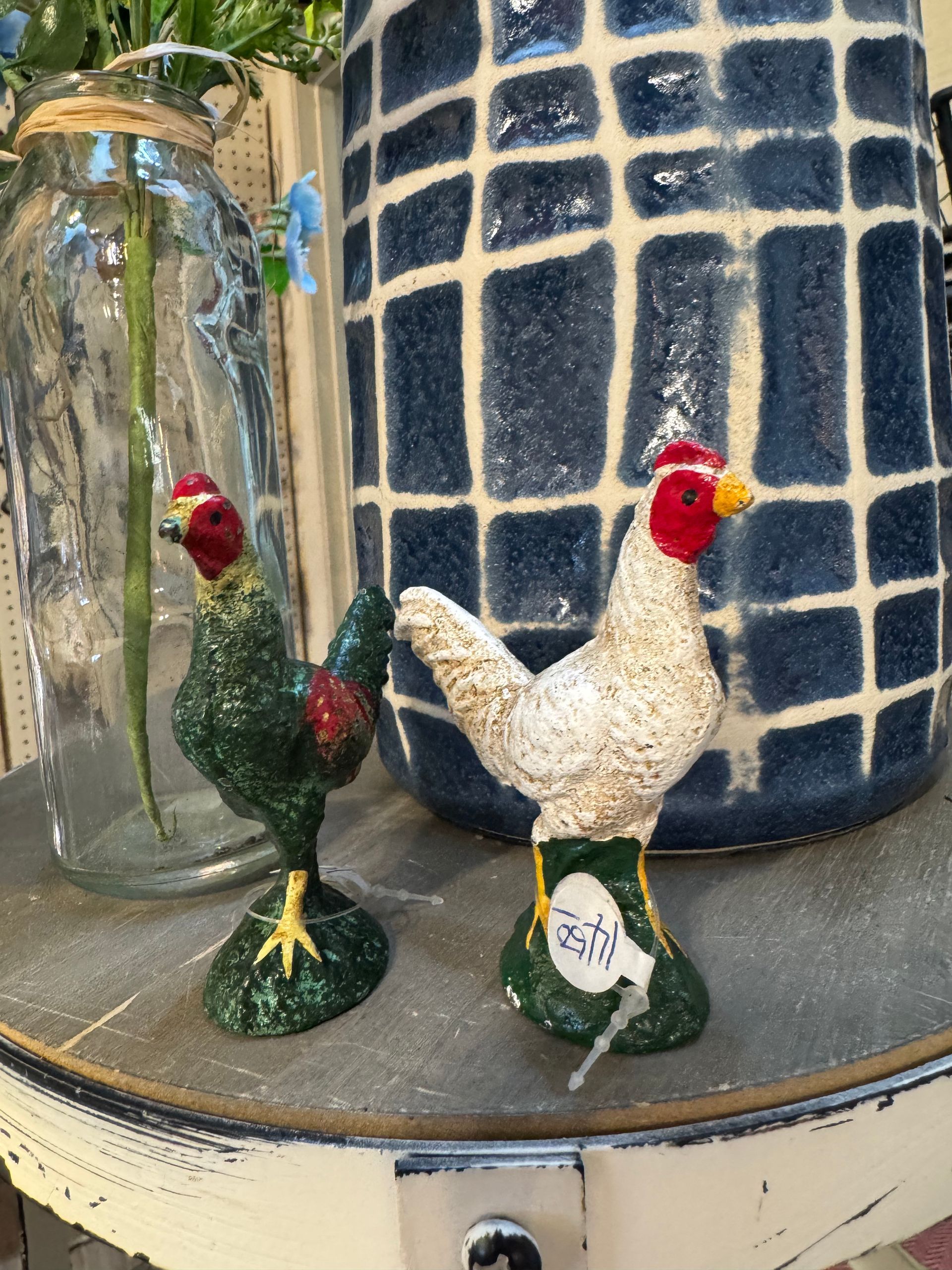 Two chicken figurines are sitting on a table next to a vase.
