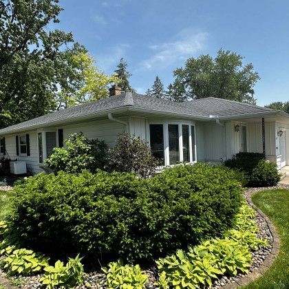 A white house with a gray roof is surrounded by bushes and trees