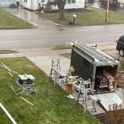 A trailer with a ladder and a table saw in the backyard of a house.