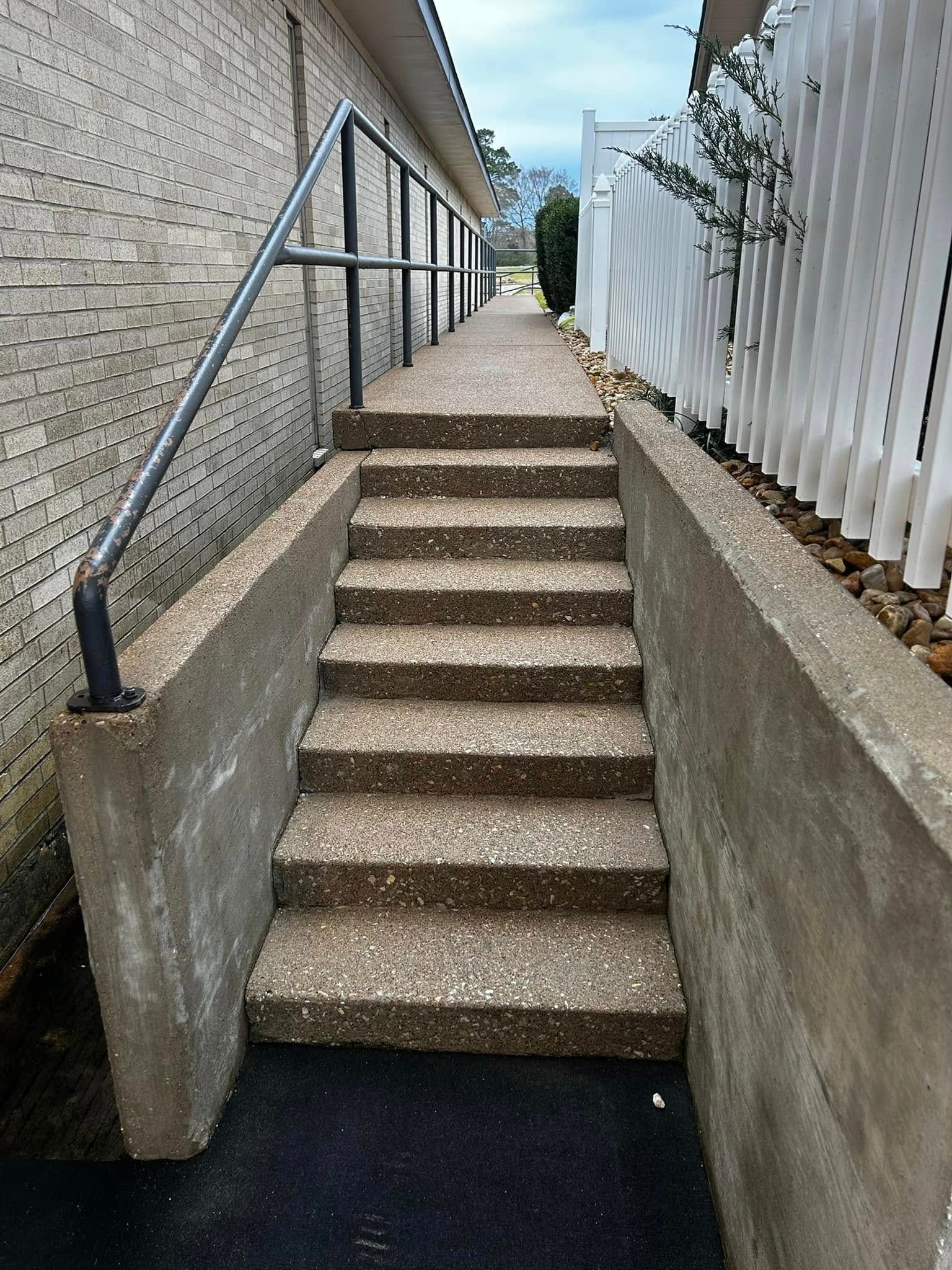 Concrete stairs leading up to a path between a brick wall and a white picket fence, with a handrail on the left.