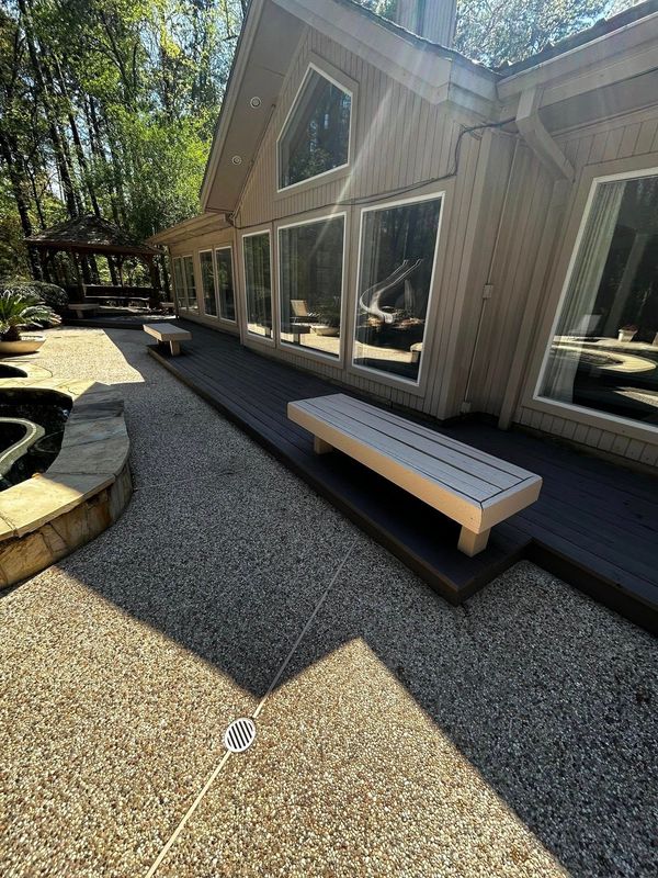 Exterior view of a house with large windows, a deck, and pebbled pathway. Wooden benches and trees are also visible.