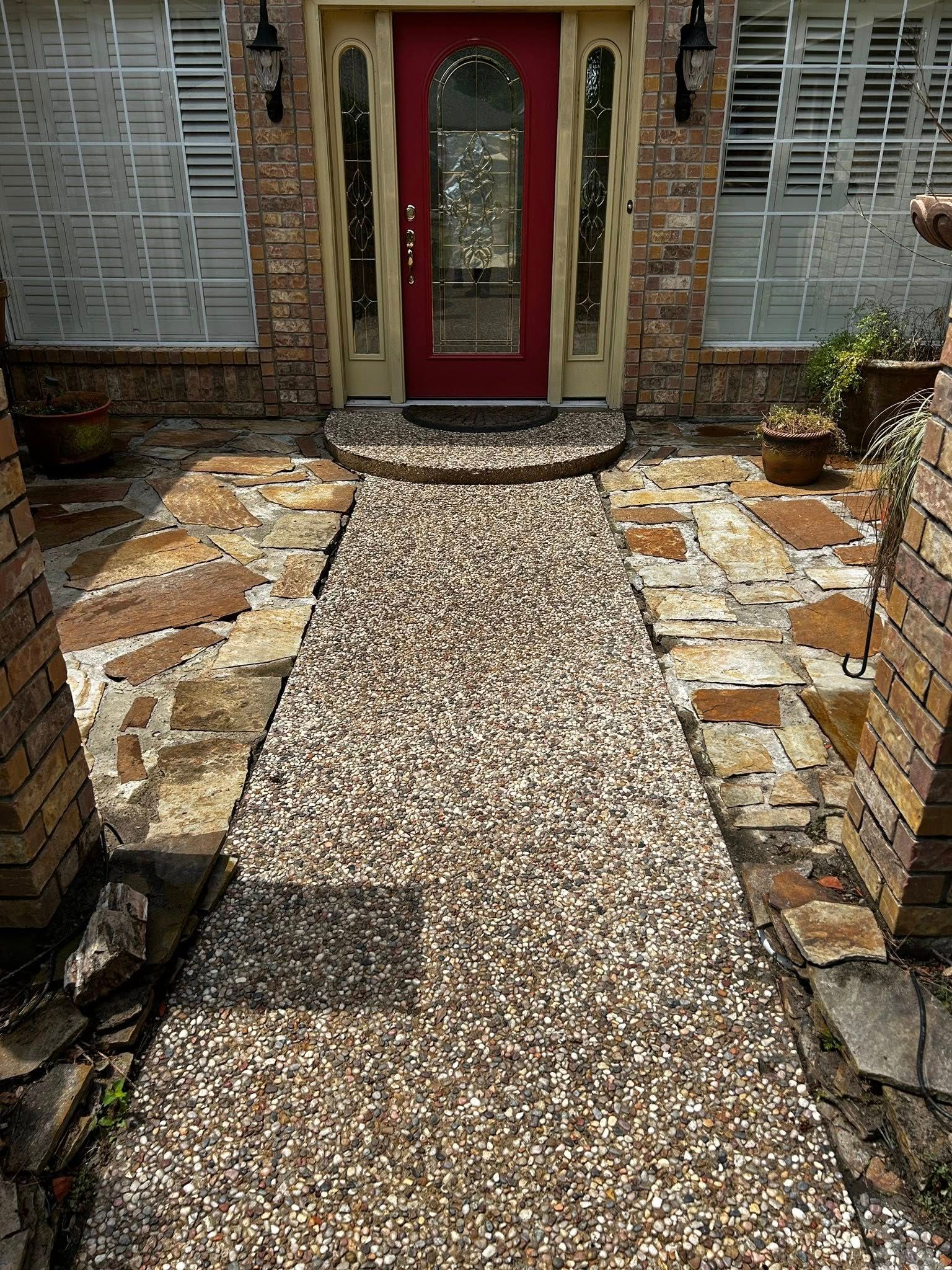 Pathway of pebble concrete leading to a red door with stone and brick exterior.