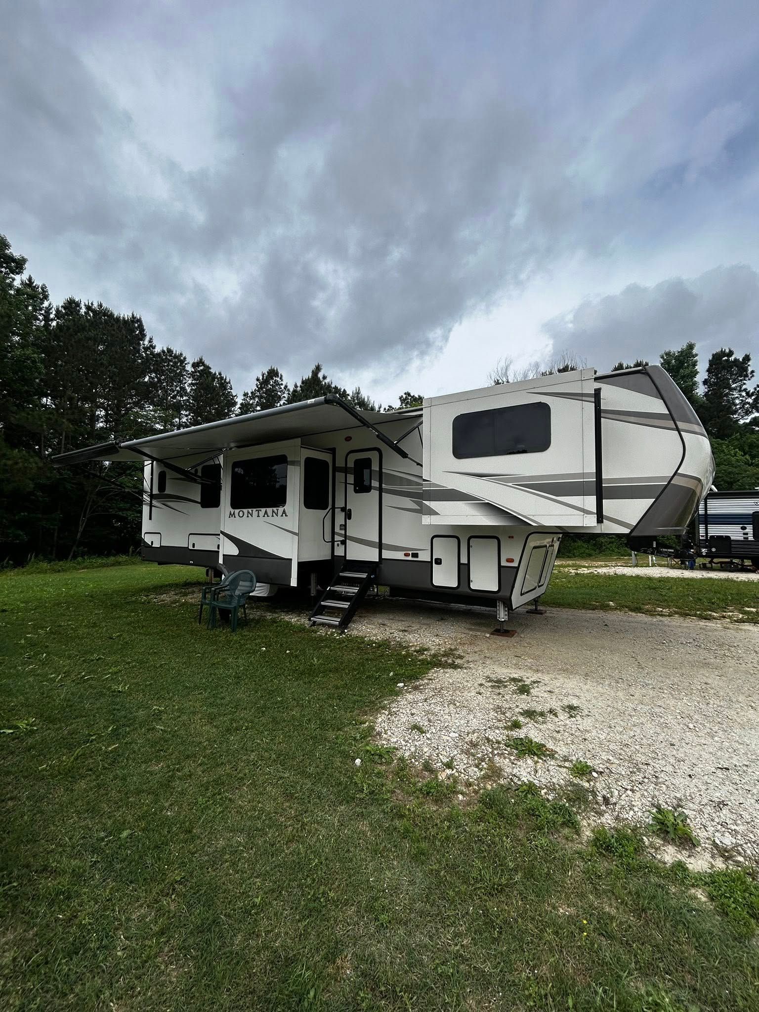 White and gray fifth-wheel camper parked on gravel and grass under a cloudy sky. Awning open.