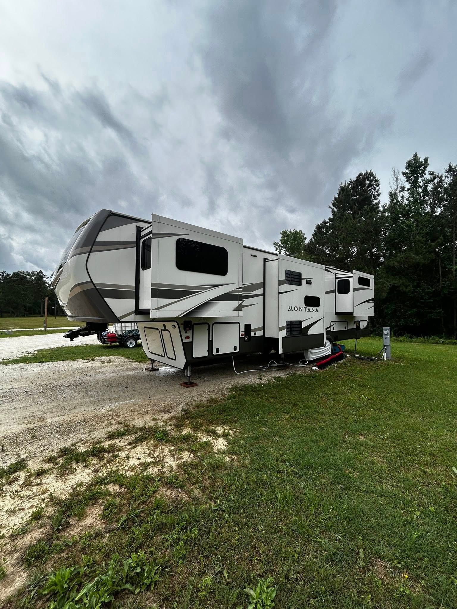 White and gray fifth-wheel camper parked on gravel and grass under cloudy sky.