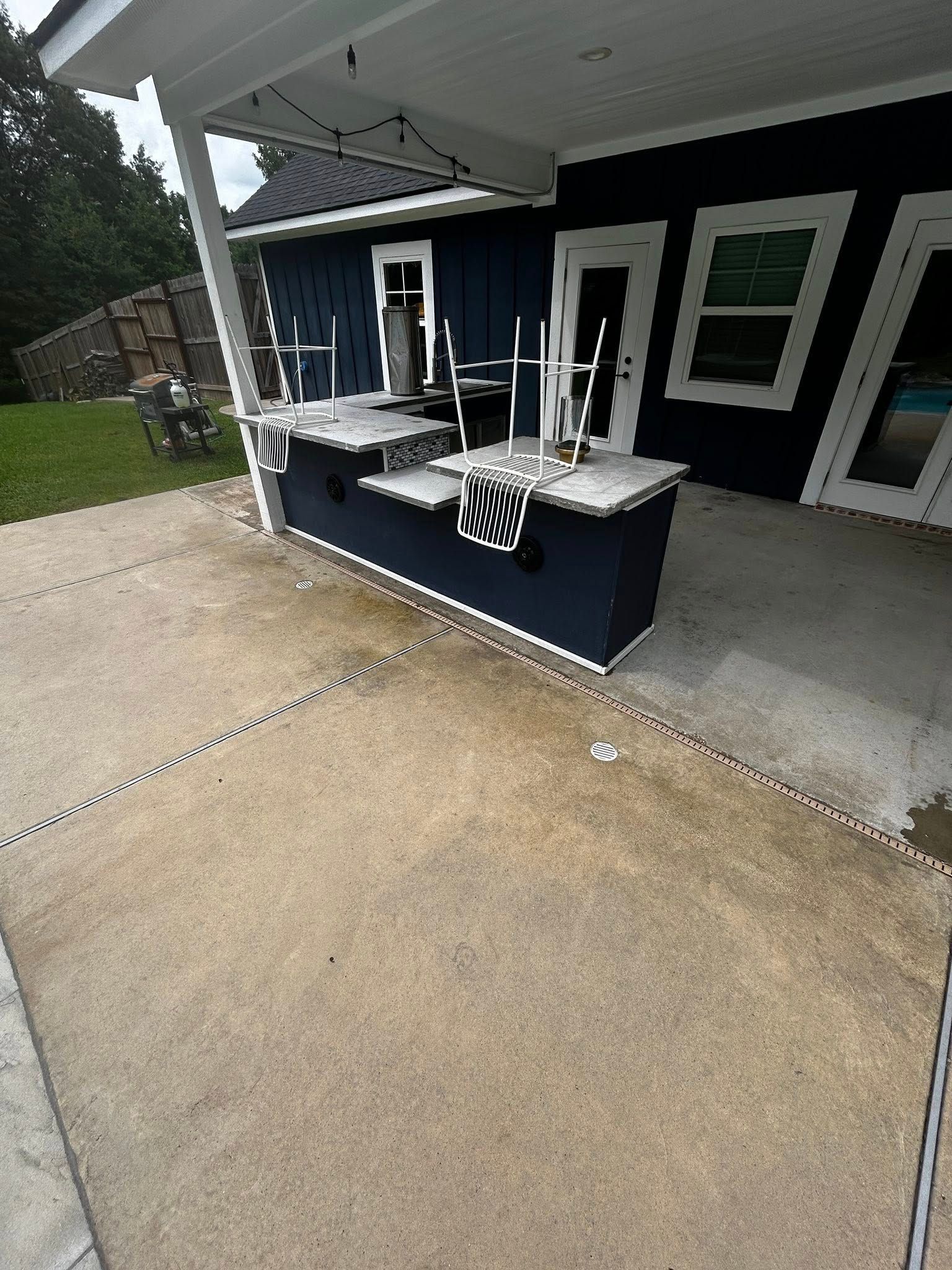 Outdoor bar with blue cabinetry, white countertops, and chairs on a concrete patio under a covered area.