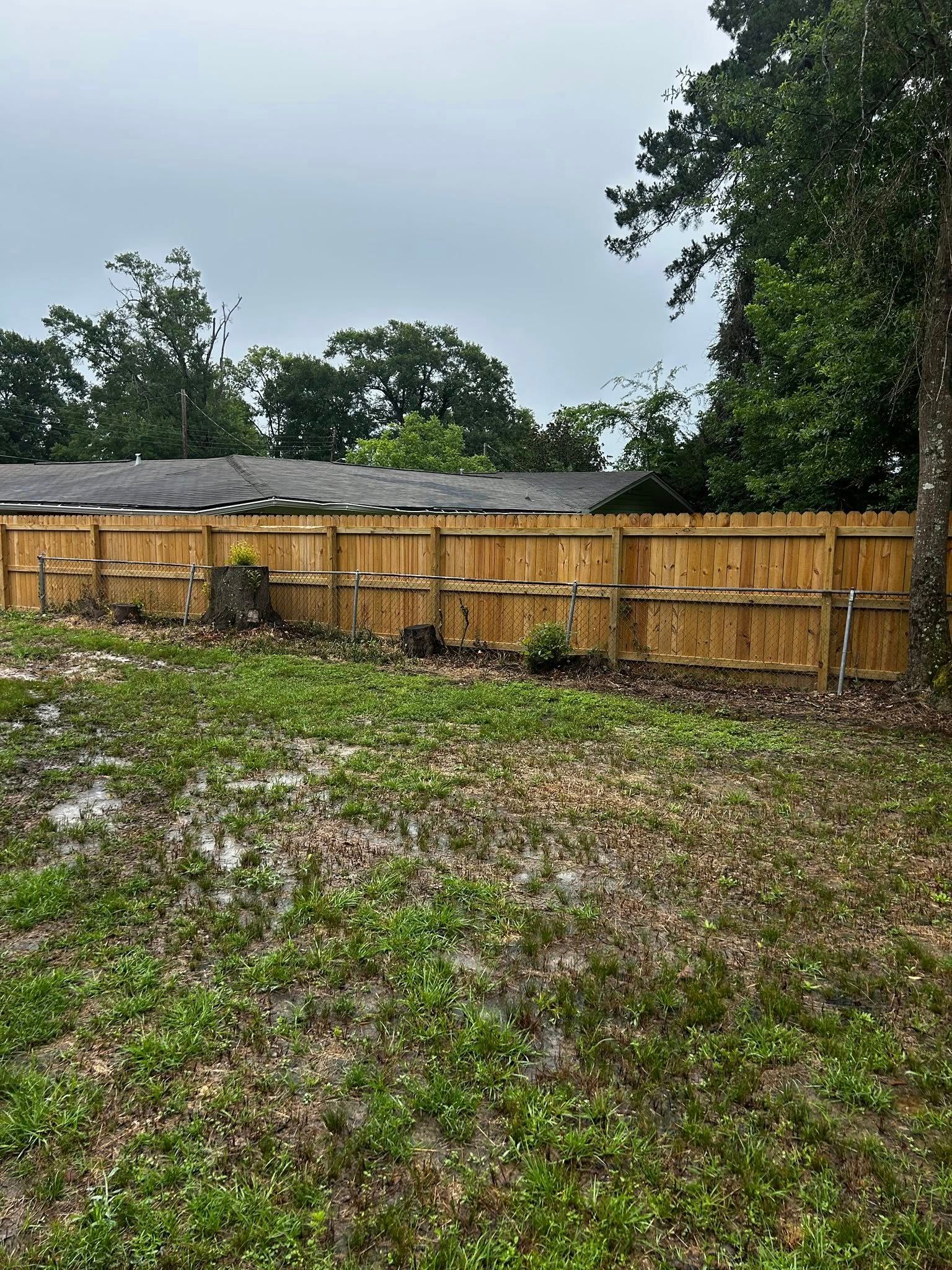 Wooden fence in a backyard with patchy grass and trees against a cloudy sky.