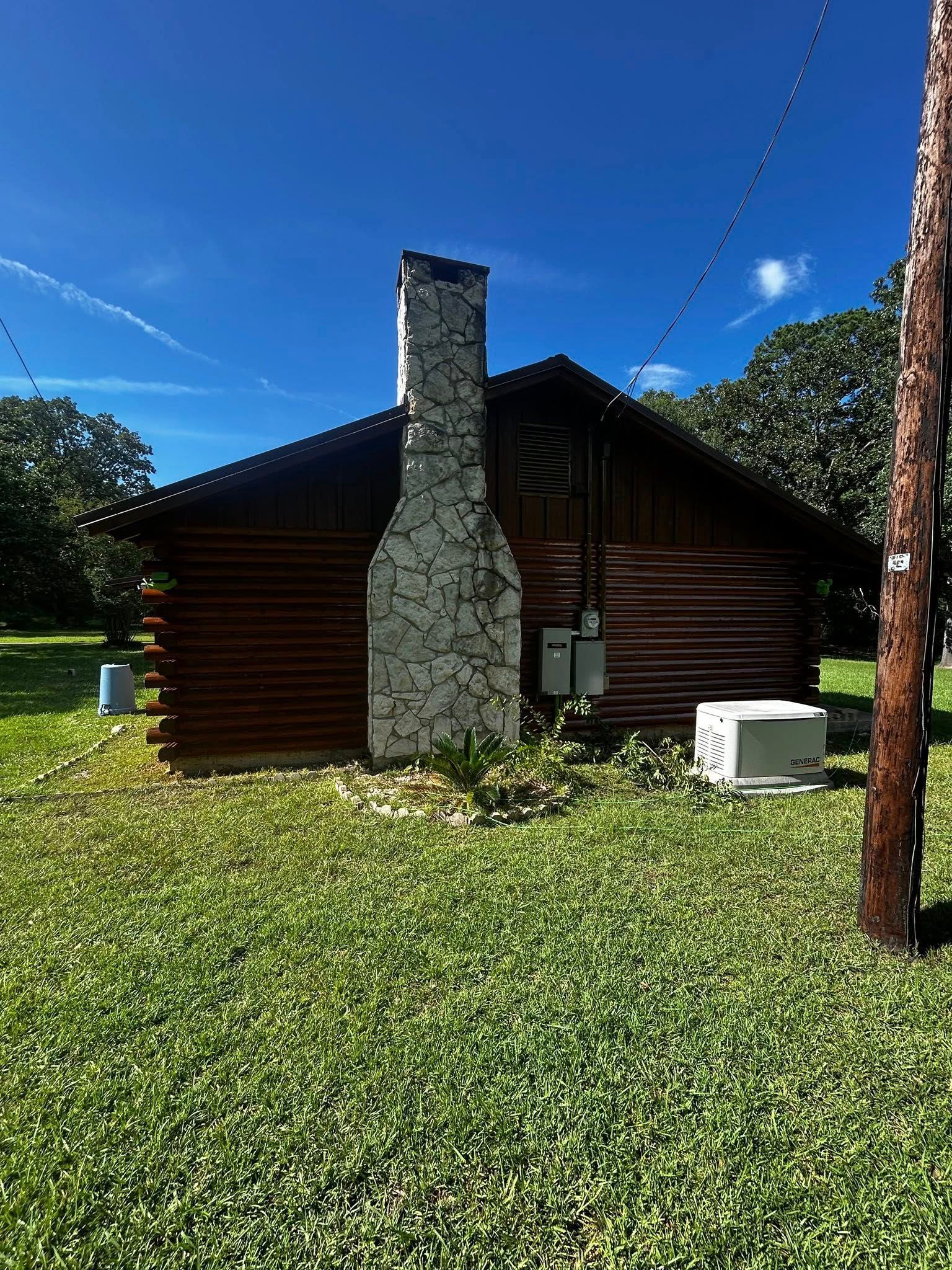 Log cabin with stone chimney, surrounded by green grass under a blue sky.