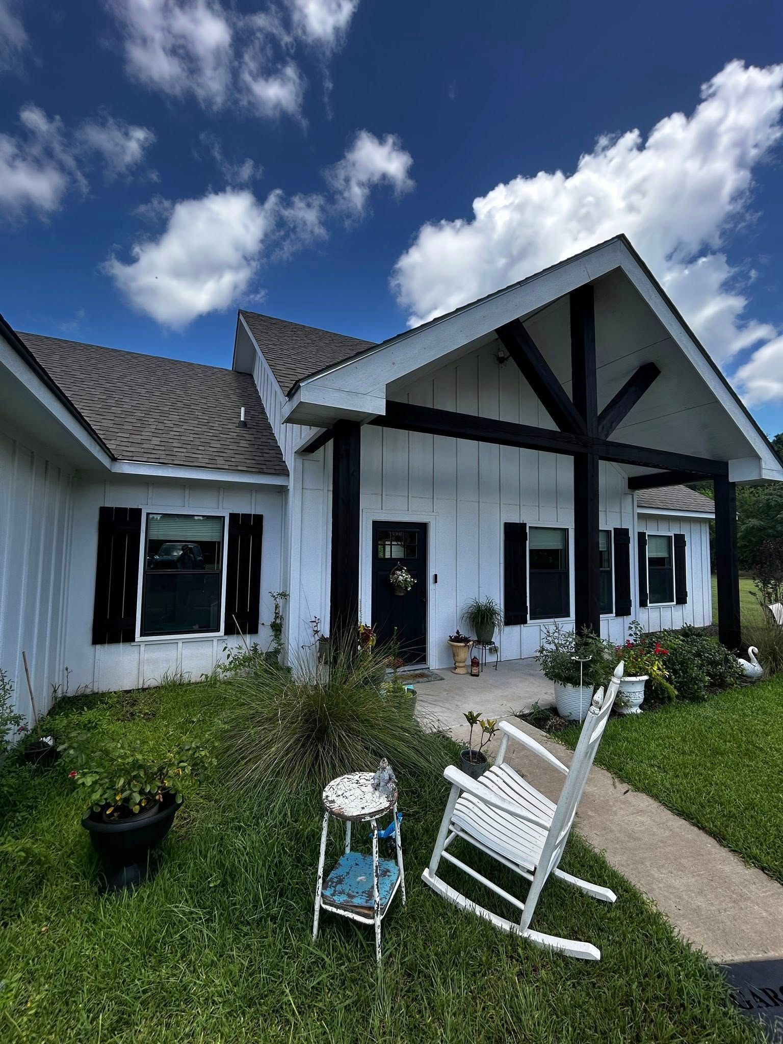 White house with black trim, rocking chair, and overgrown yard under a blue sky with clouds.