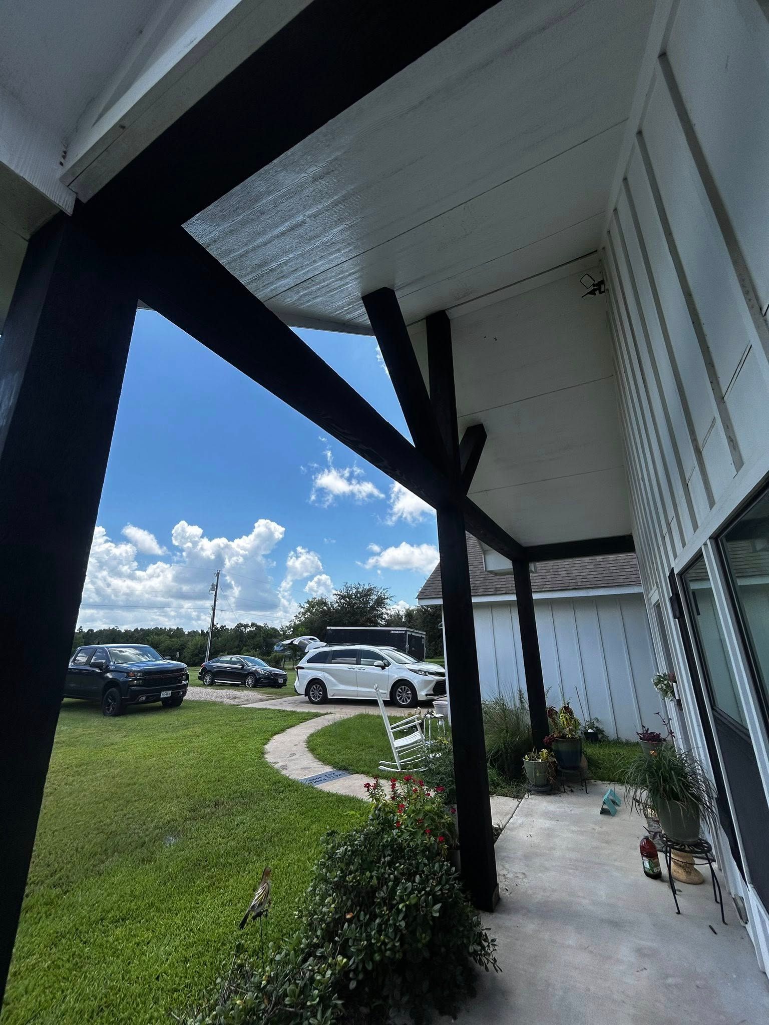 Exterior view of a house's porch with black beams, white ceiling, and parked cars in the yard.