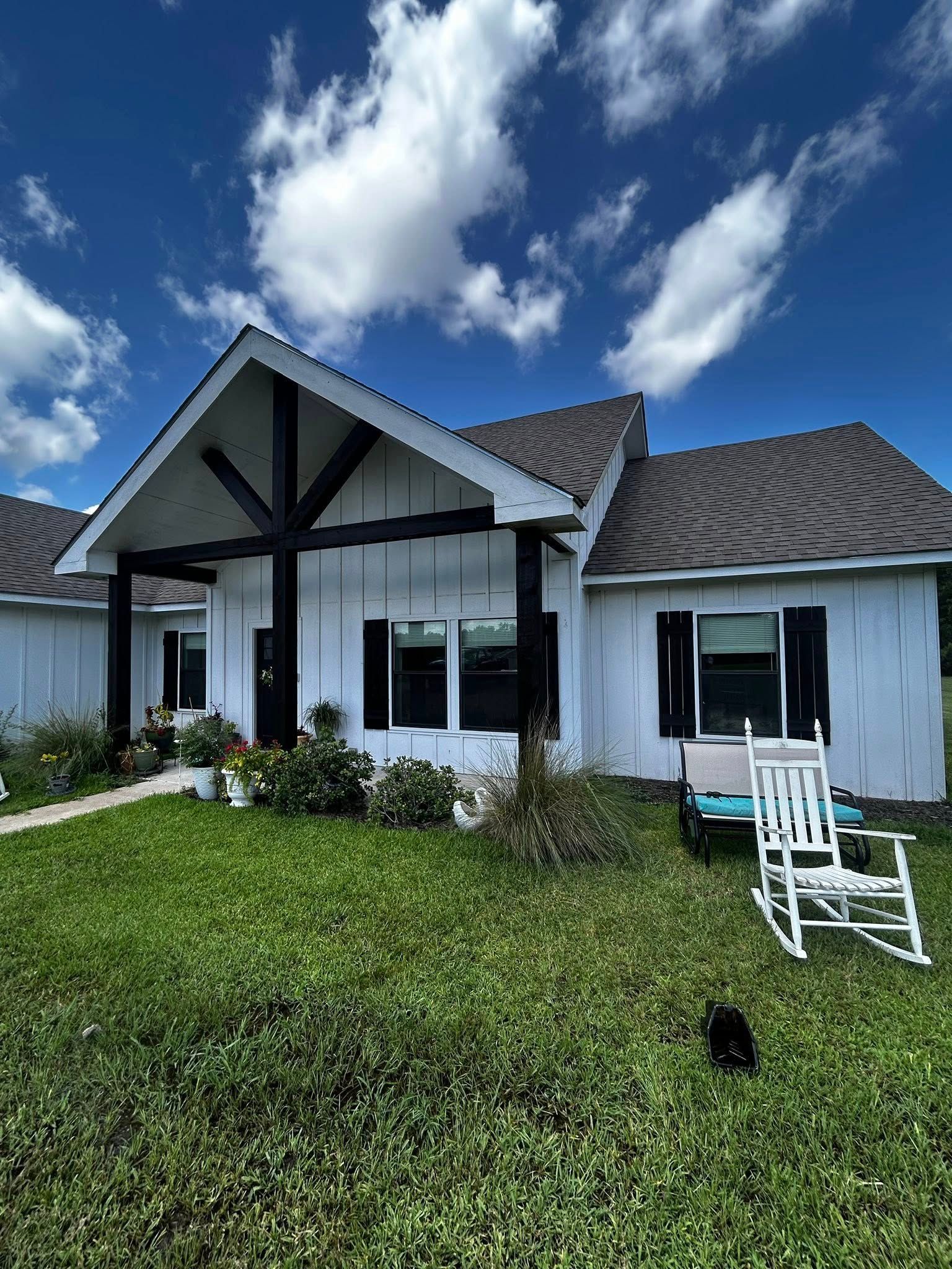 White house with black accents, porch, rocking chair on the lawn, bright blue sky with fluffy clouds.