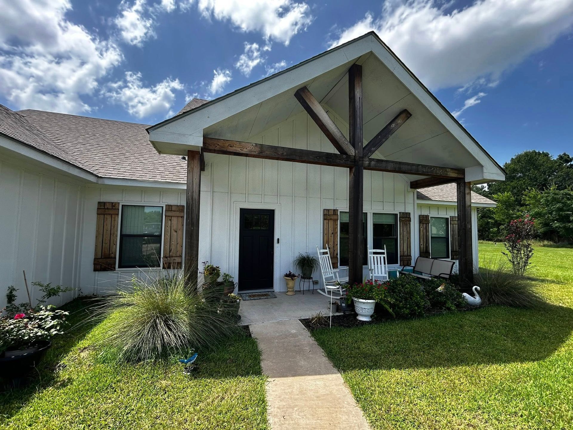 White house with porch; wooden beams and shutters. Path leads to black door. Cloudy sky, green grass.