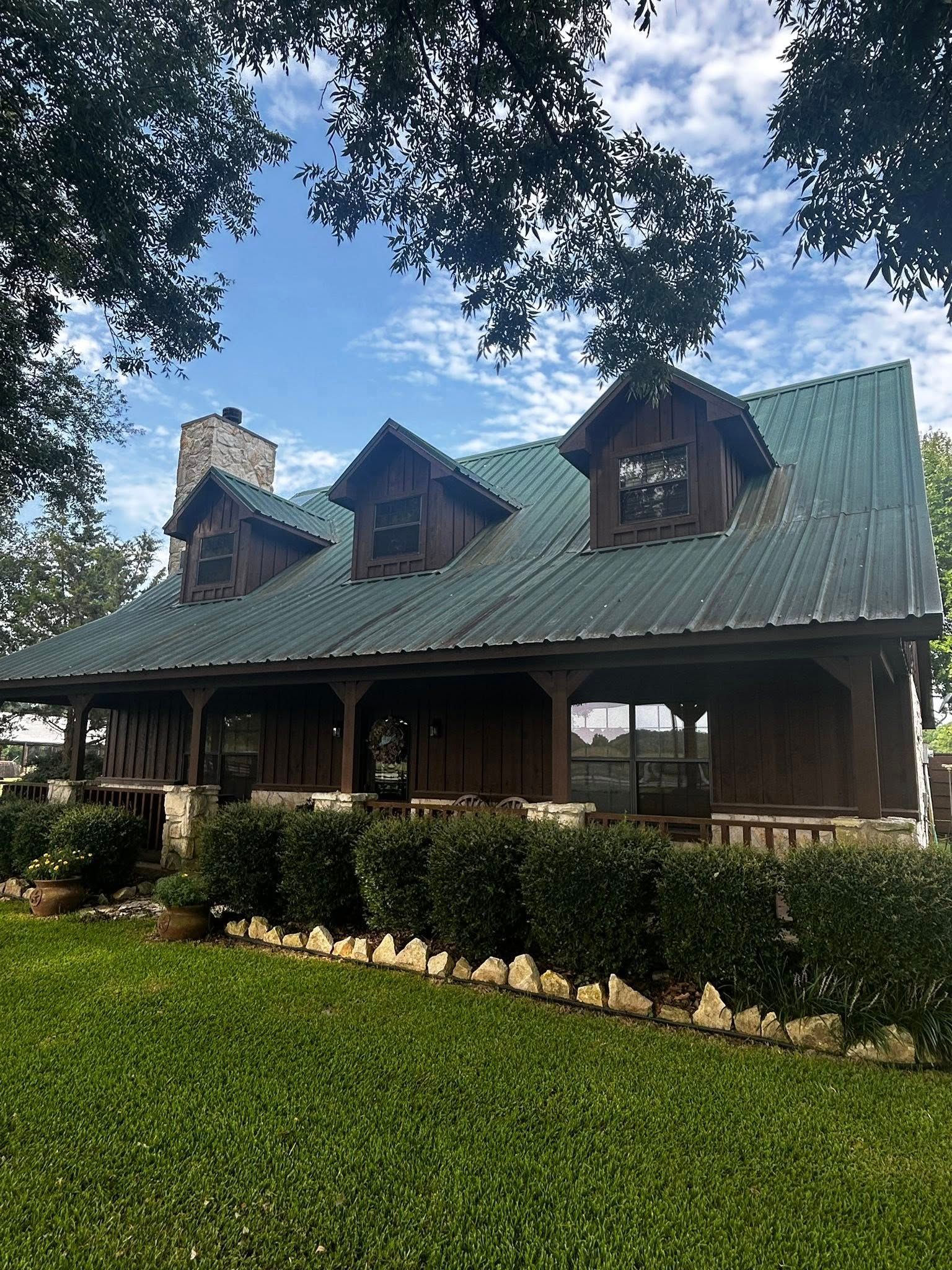 Brown cabin-style house with green metal roof, three dormers, stone chimney, and covered porch, set on a green lawn.