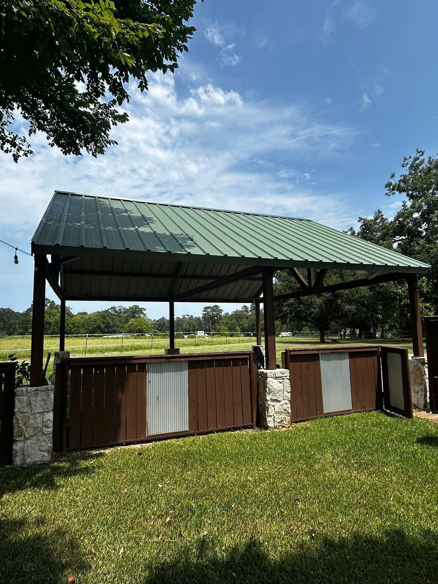 Outdoor shelter with green metal roof, brown wooden walls, and stone pillars, in a grassy area under a blue sky.