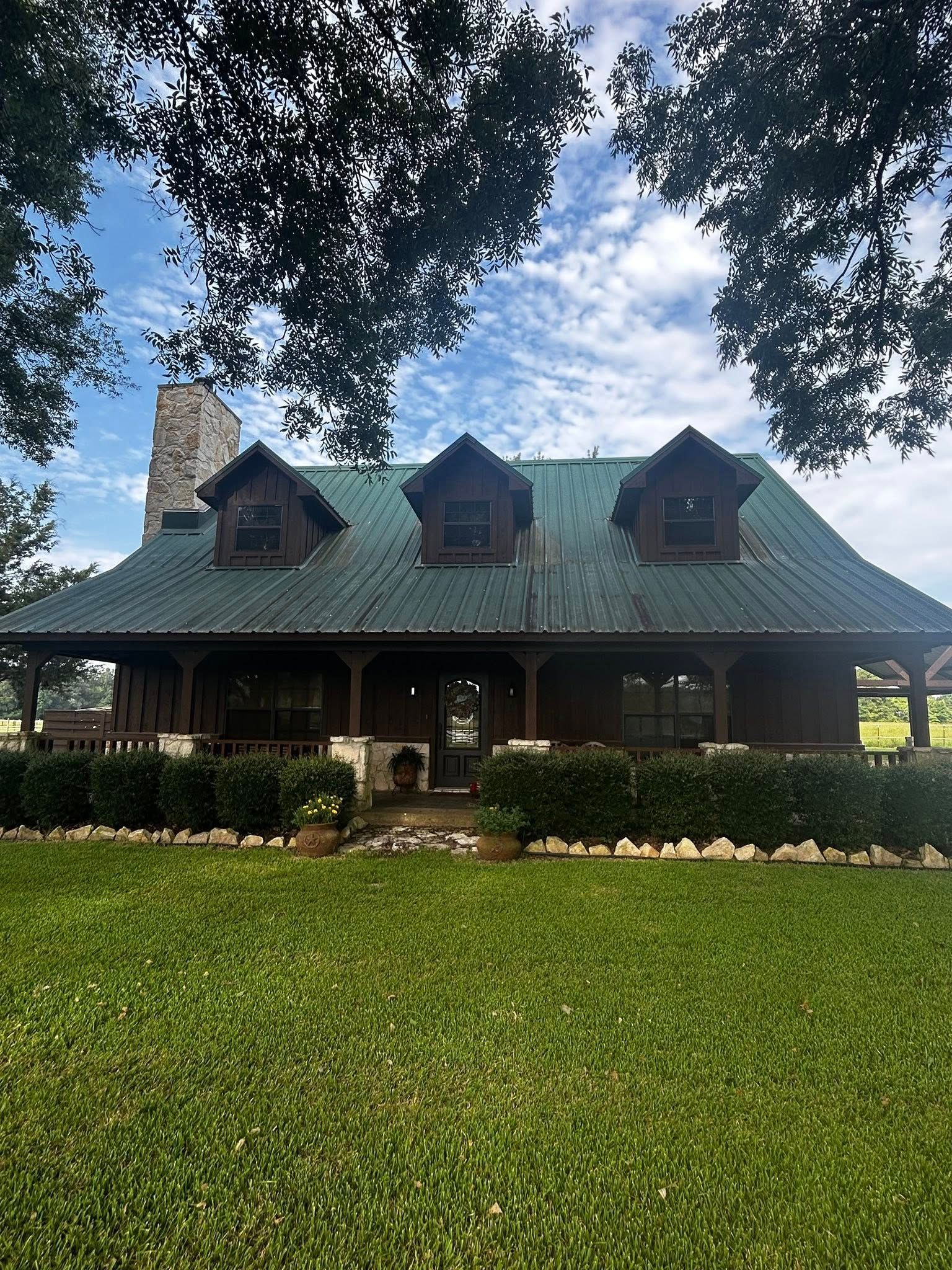 A brown house with a green roof, three dormers, and a stone chimney, set on a green lawn with a cloudy sky.