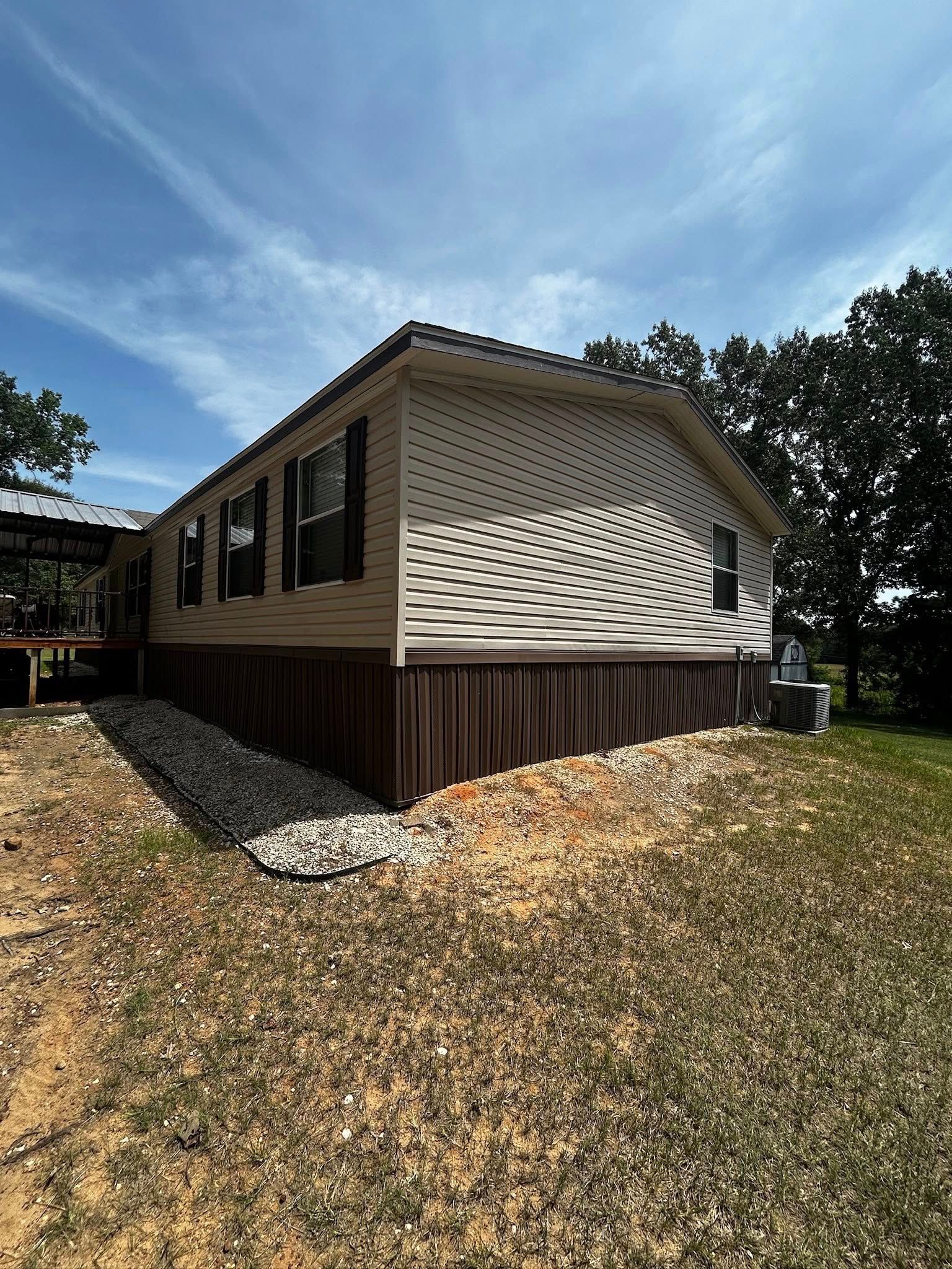 Side view of a beige and brown manufactured home with black shutters, on a grassy lot under a blue sky.