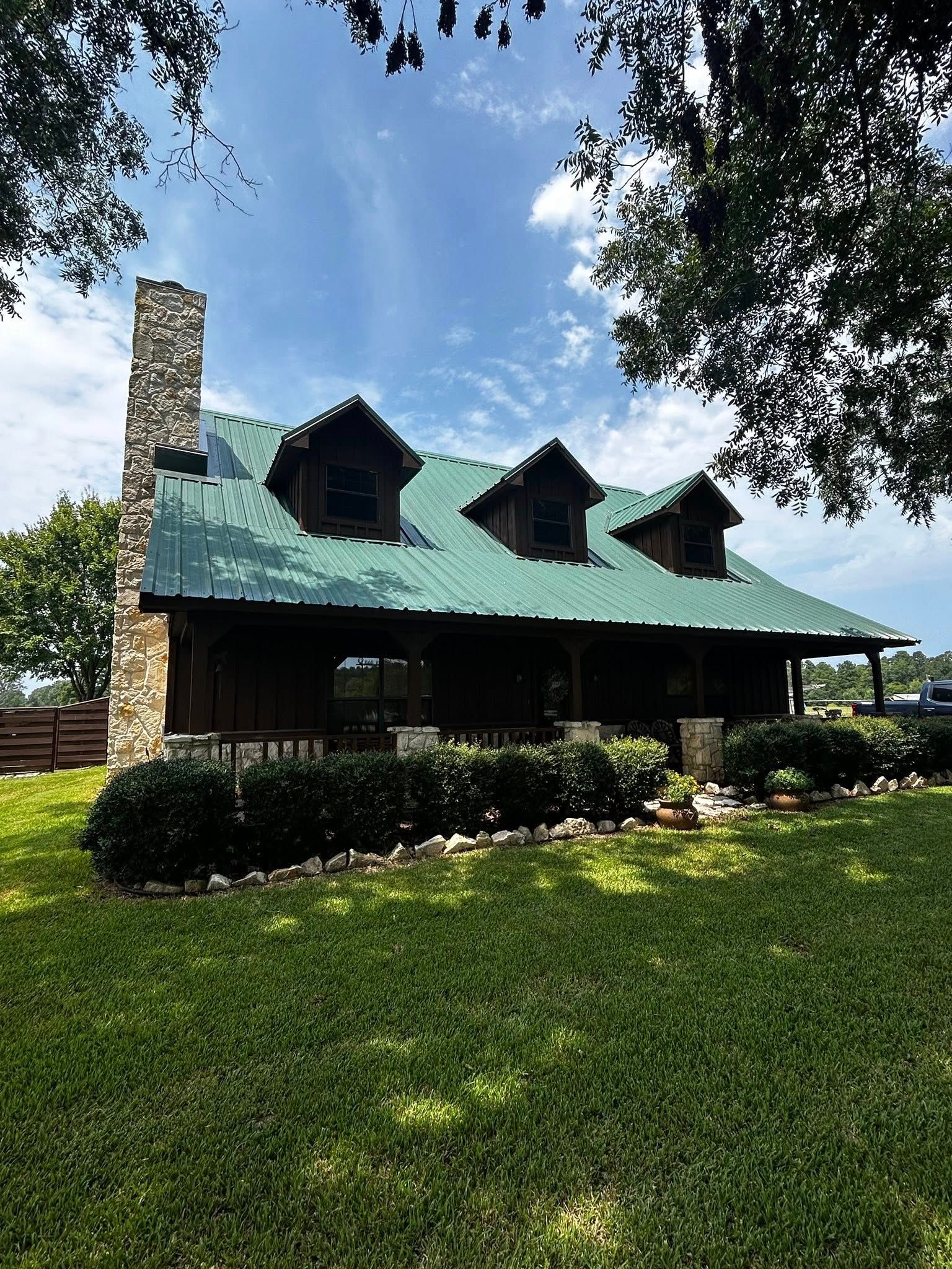 Cabin with green metal roof, stone chimney, and three dormers, surrounded by green grass and trees.