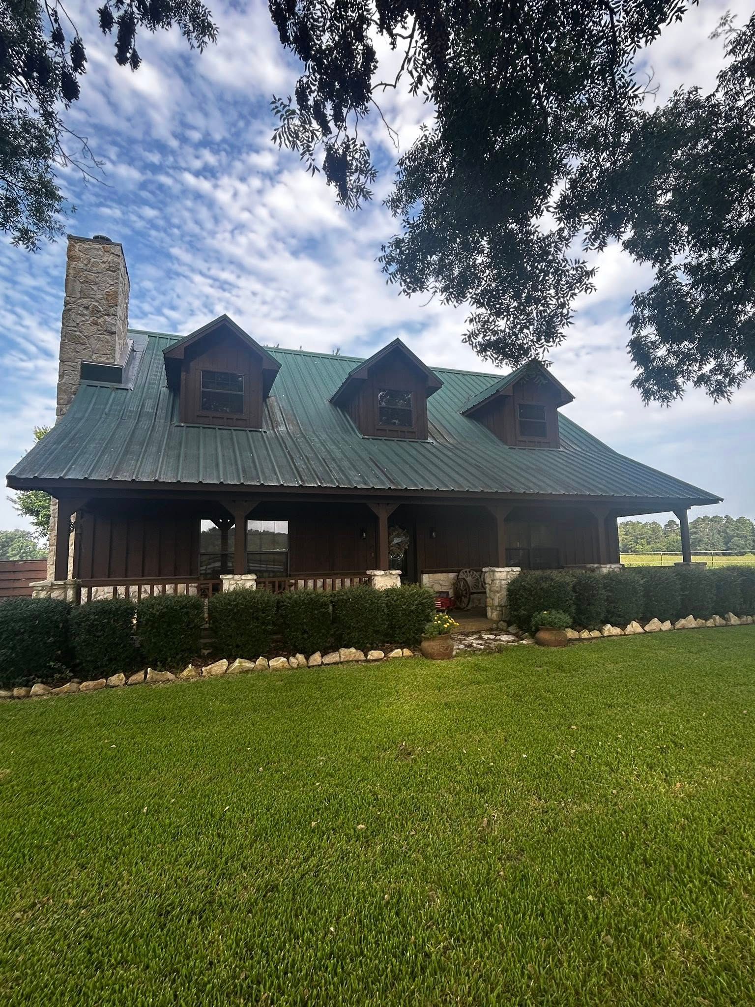 Rustic brown cabin with green roof, stone chimney, three dormers, and porch, surrounded by green lawn and trees.
