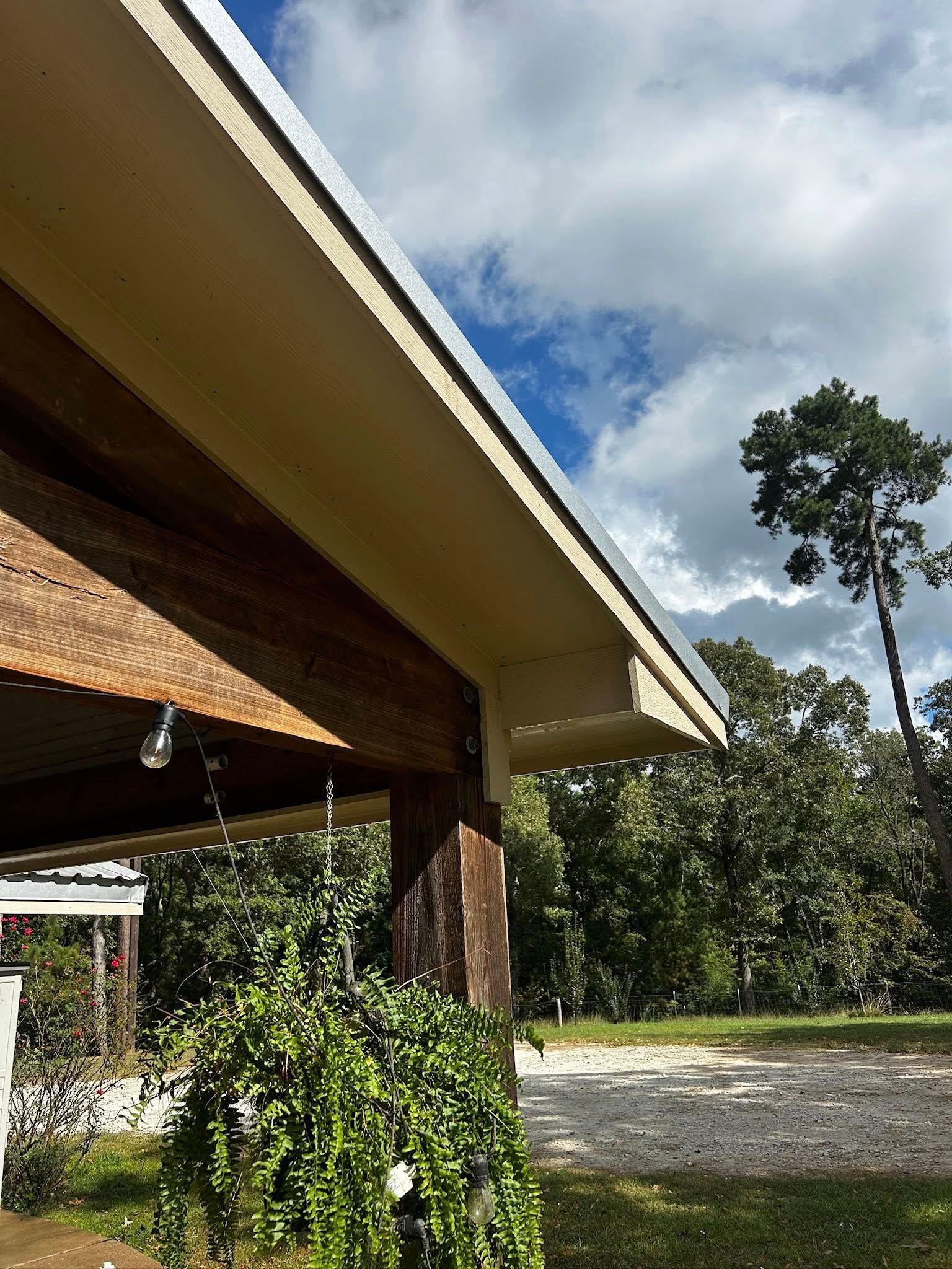 Overhanging roof with a gutter, supported by wooden beams. Green foliage, trees, and sky in the background.