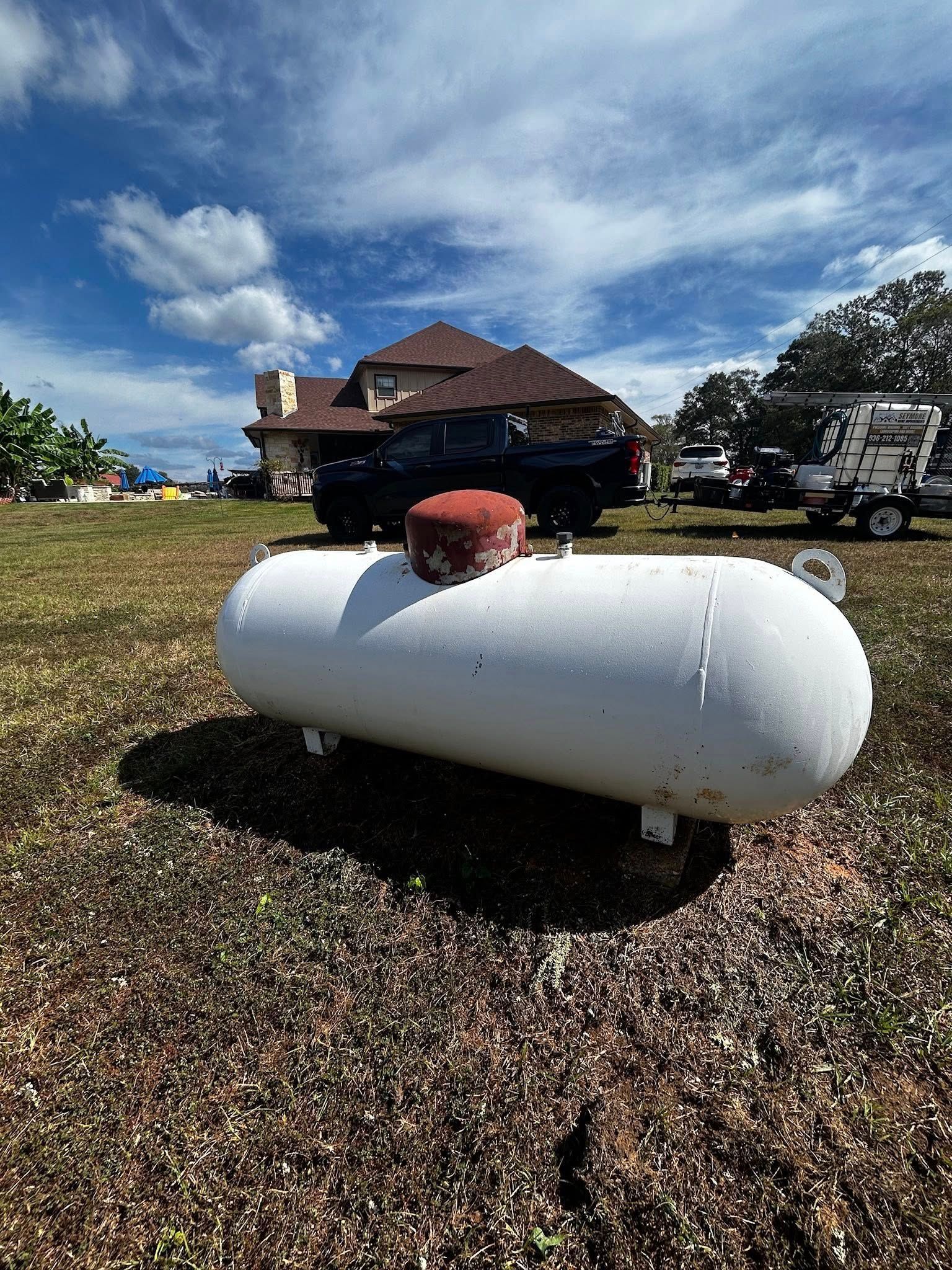 White propane tank in a grassy field with a house in the background on a partly cloudy day.