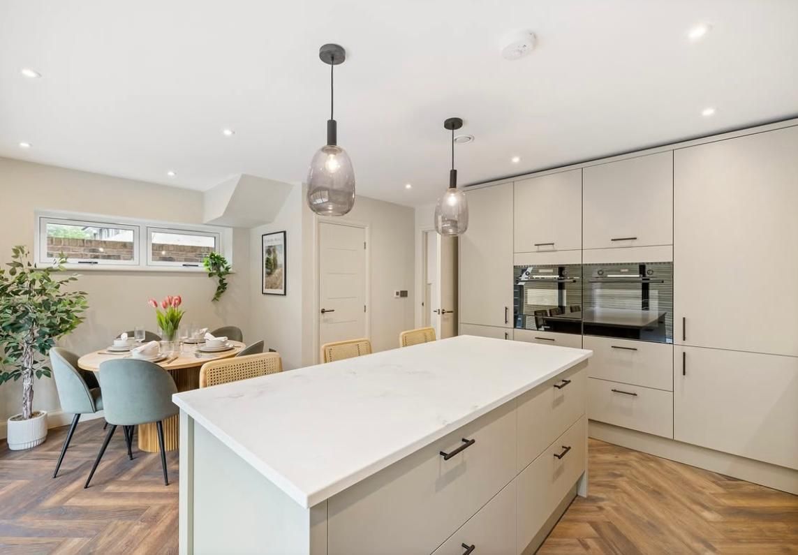 A modern kitchen with a white island, beige cabinets, pendant lighting, and a dining area with a round table and chairs.