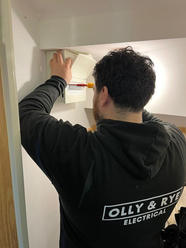 An electrician in a black hooded sweatshirt works on a white consumer unit mounted on a wall.