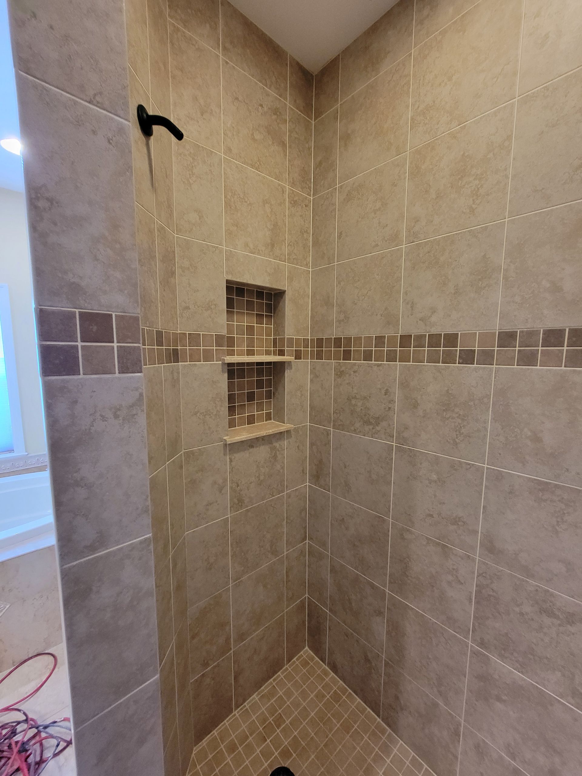Tiled shower with corner seat. Beige tiles with a darker stripe. Two recessed shelves. Black shower head.