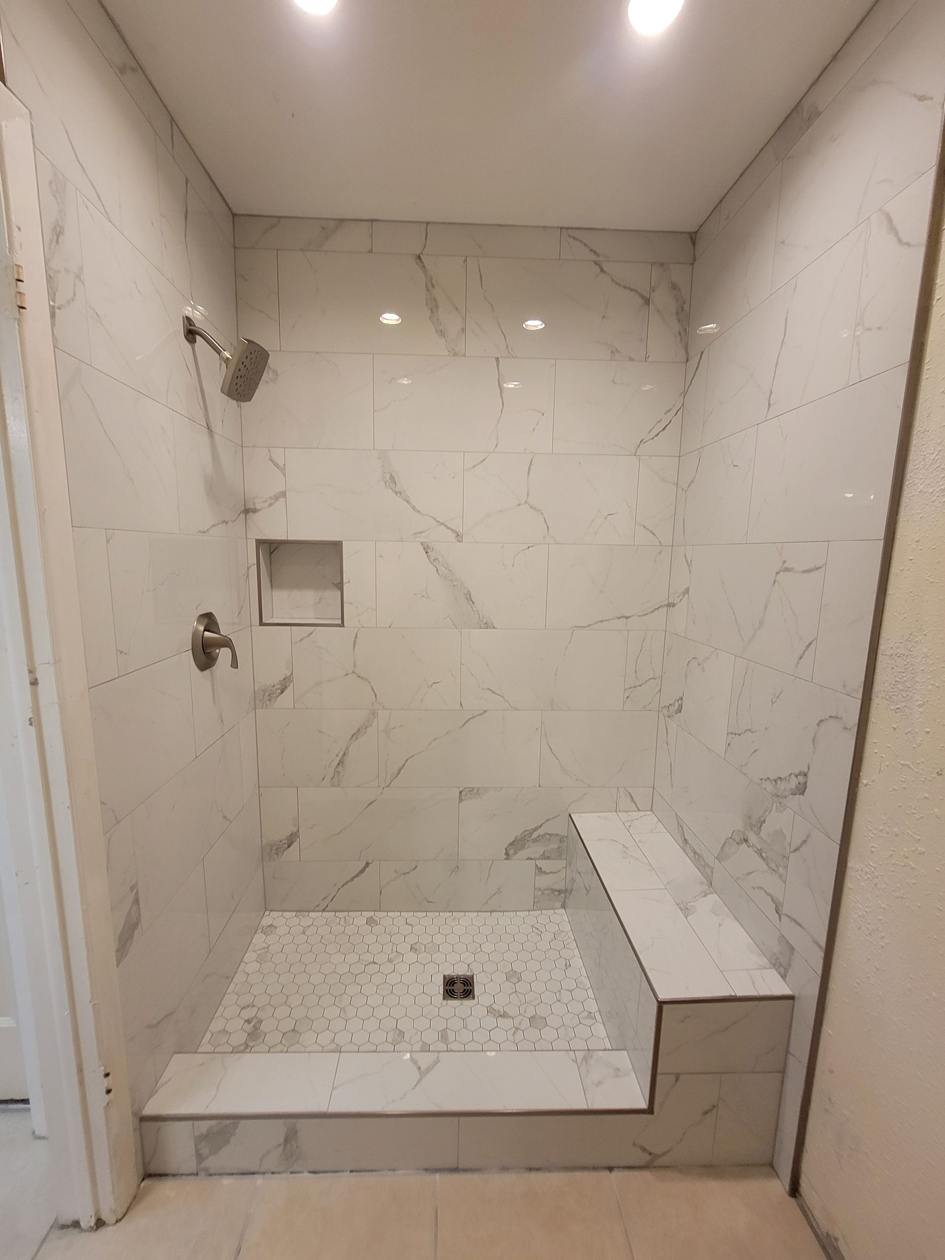 Tiled shower stall with a built-in bench. White marble-look tiles, showerhead, and a recessed soap niche.