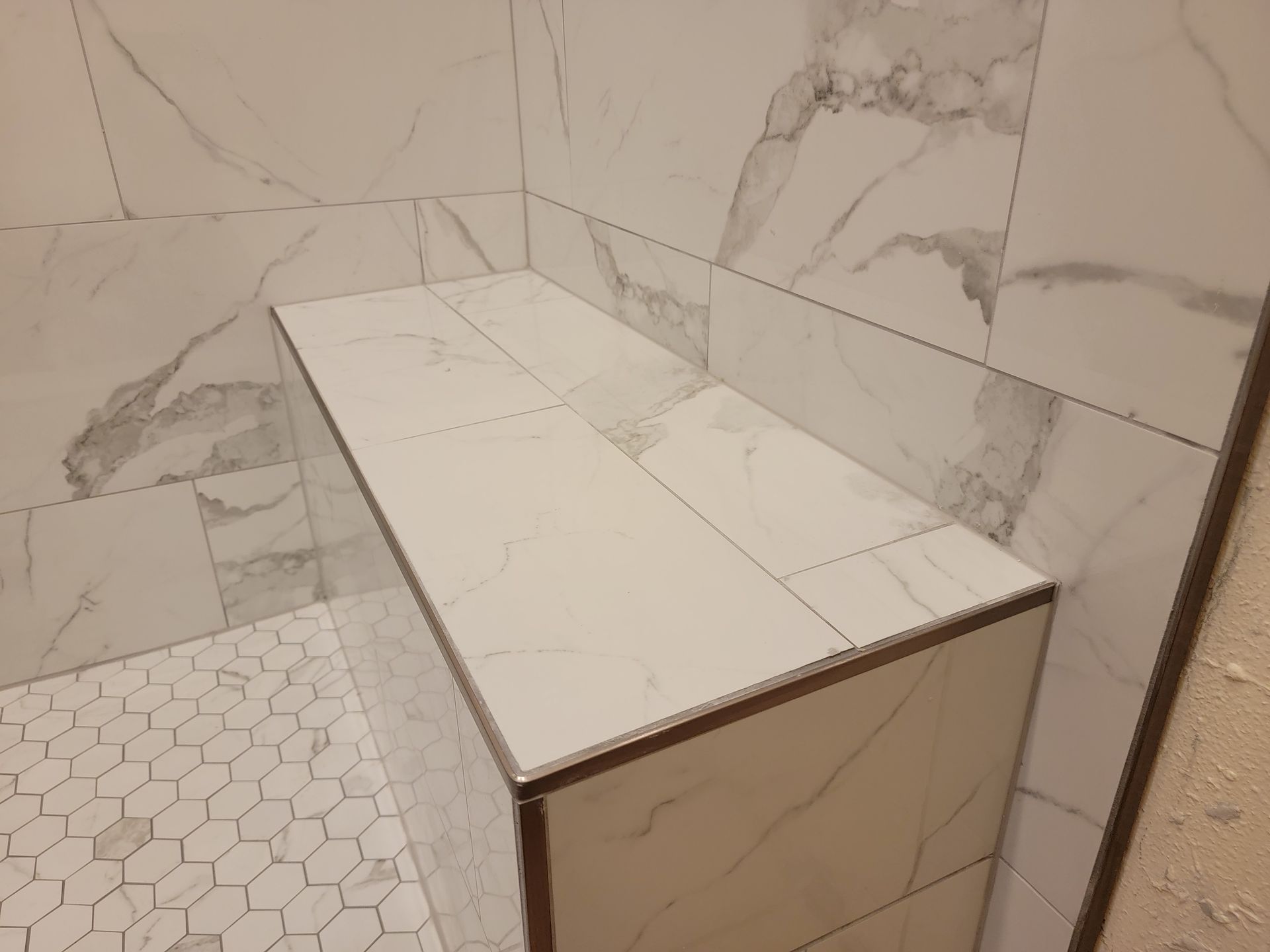 Tiled shower bench with white marble-patterned tiles; a corner shower. Hexagon tile floor.