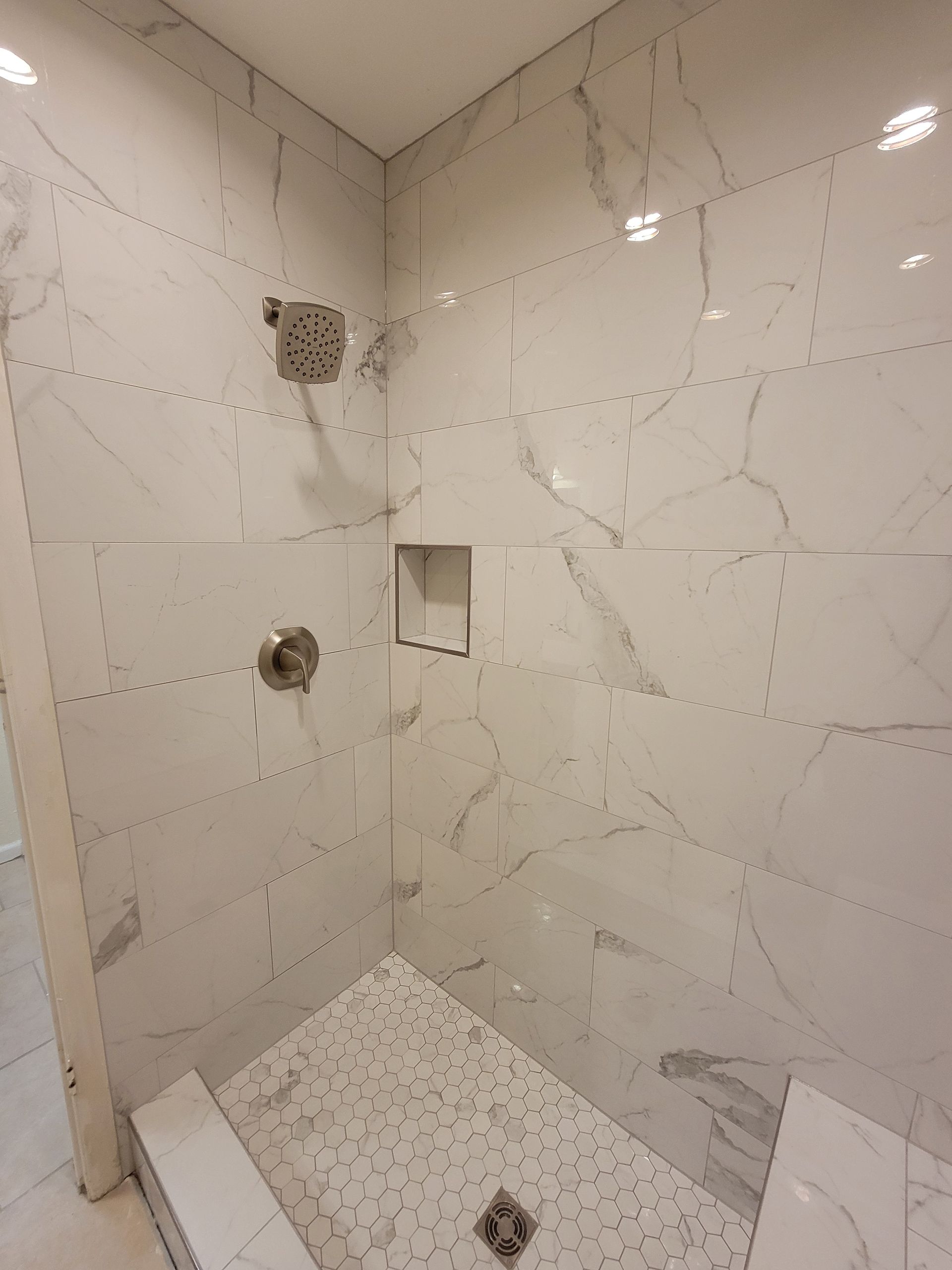 White tiled shower with marble-like veining, built-in shelf, silver shower head and controls, and a mosaic floor.