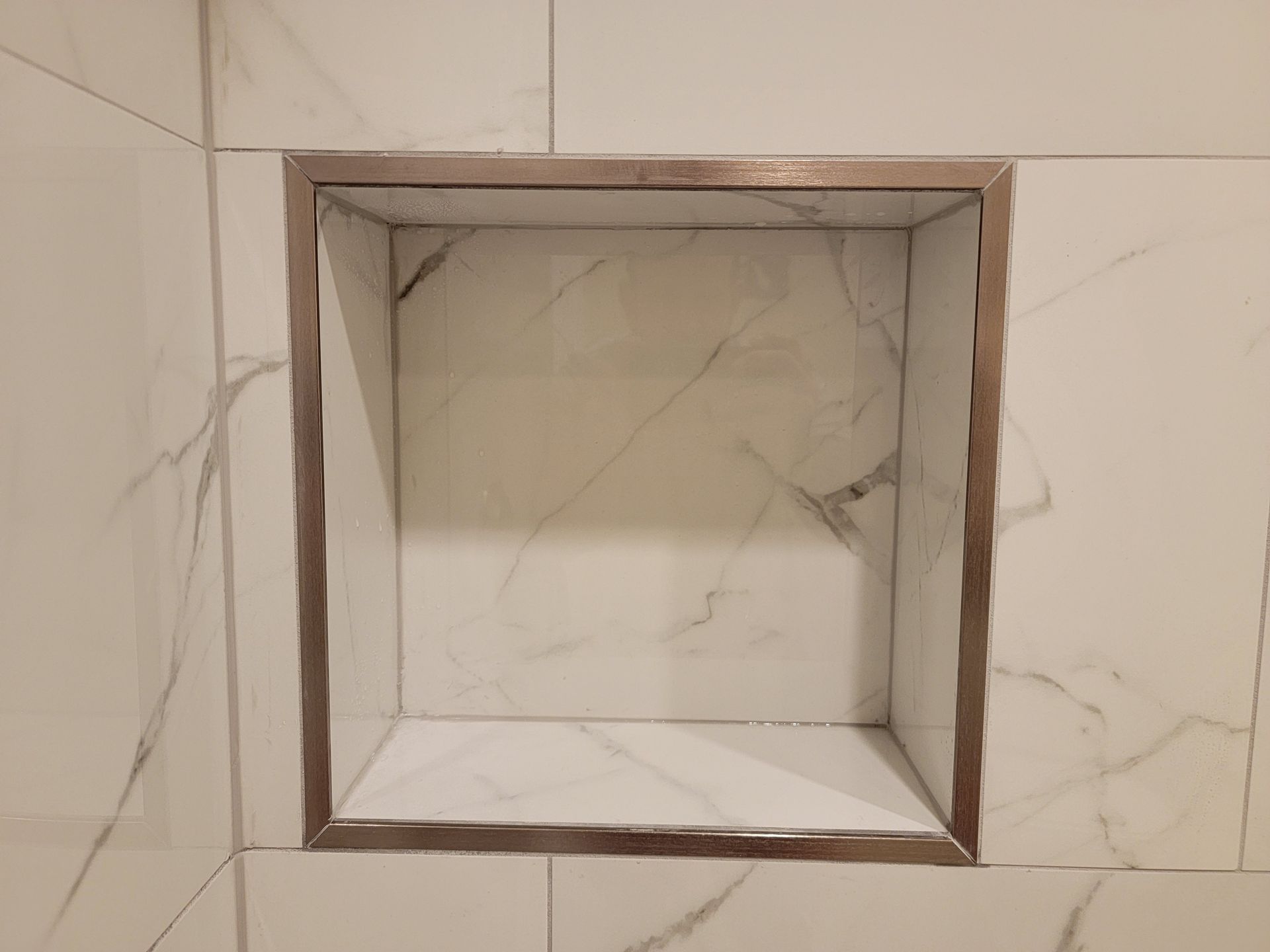 Built-in shower niche with marble-look tile, outlined in metallic trim, set into a white tile shower wall.