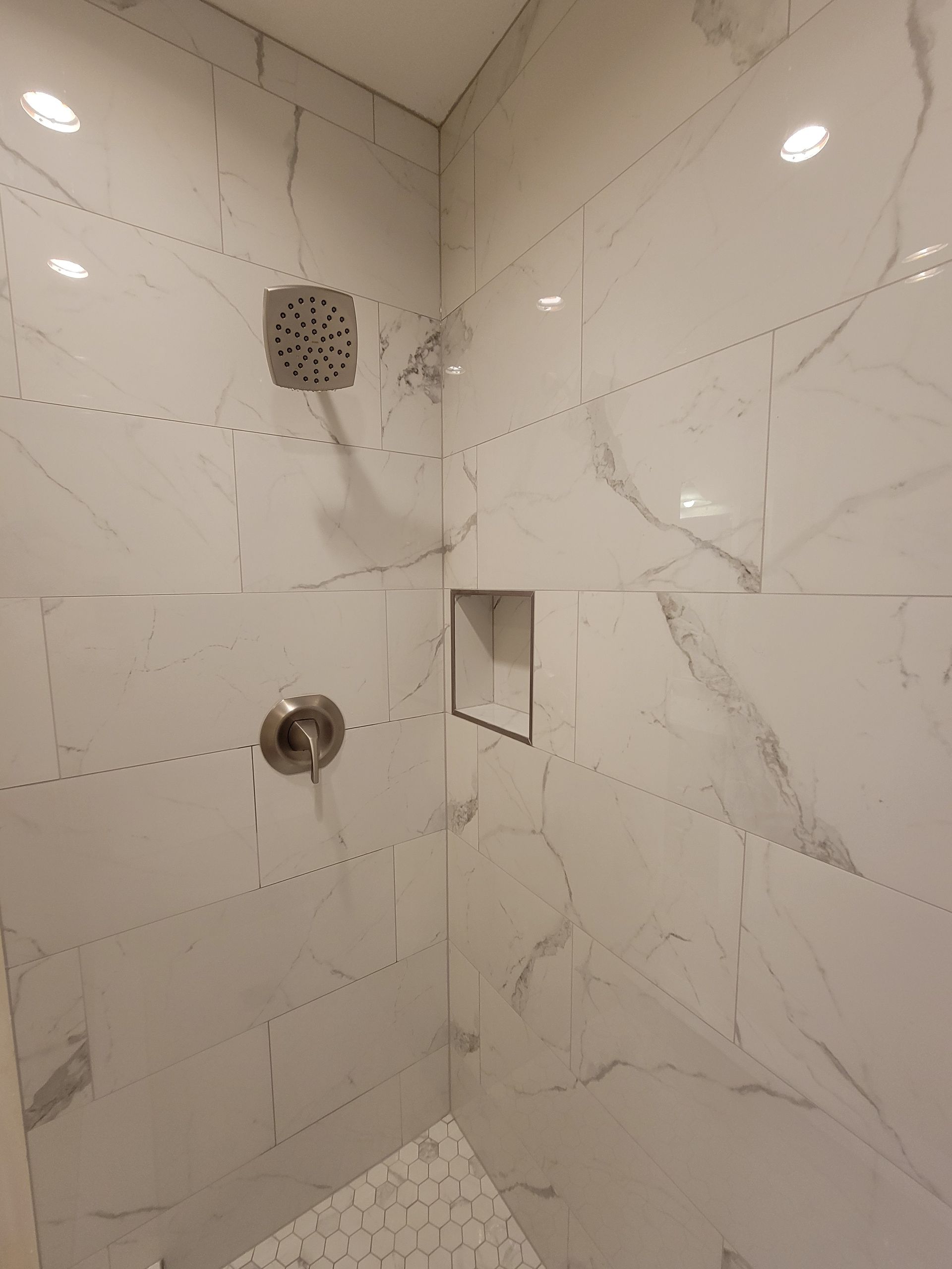 White tiled shower with a showerhead, faucet, and recessed shelf.