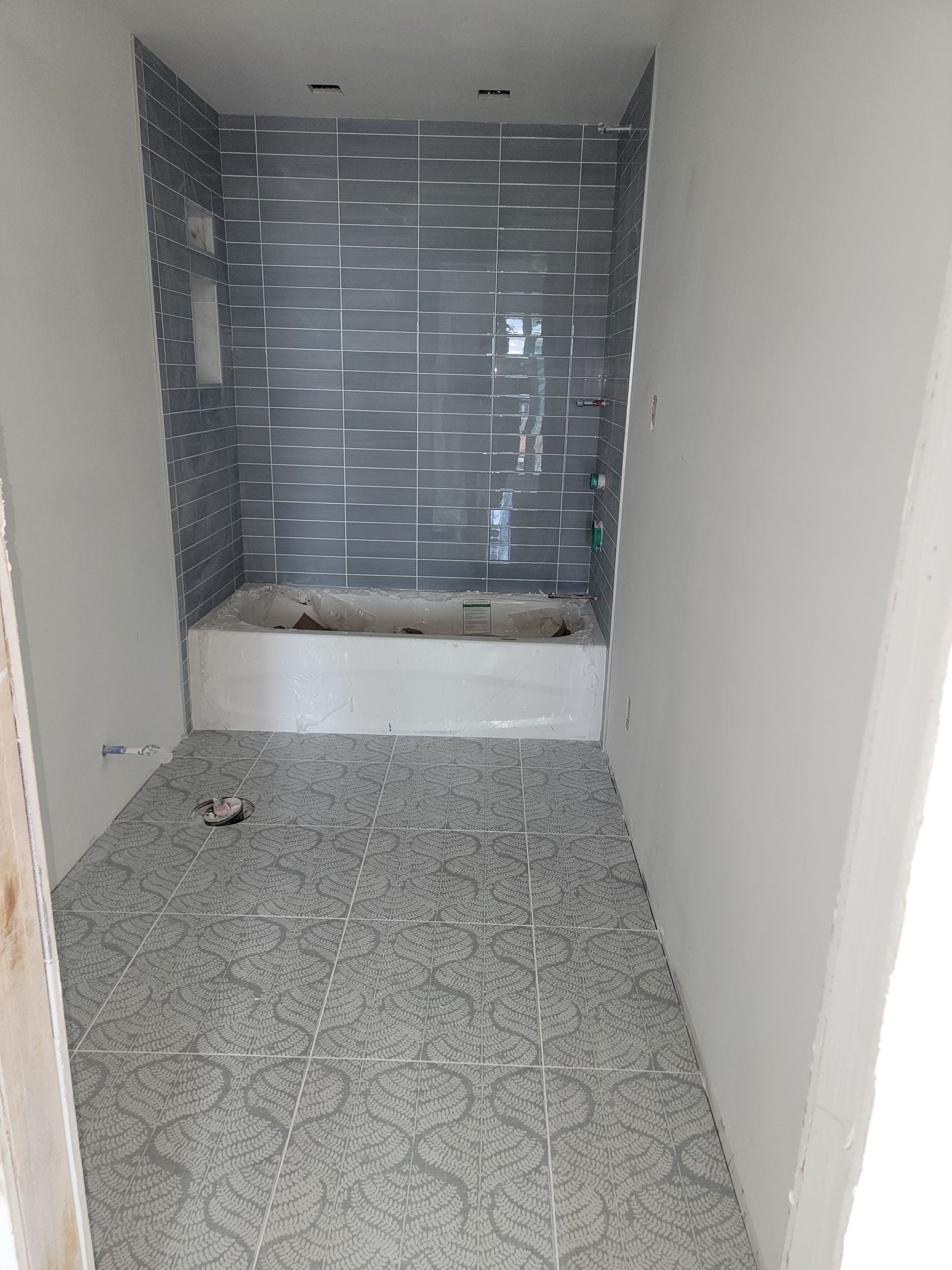 Bathroom under construction with gray patterned floor tile and blue wall tile around a white tub.