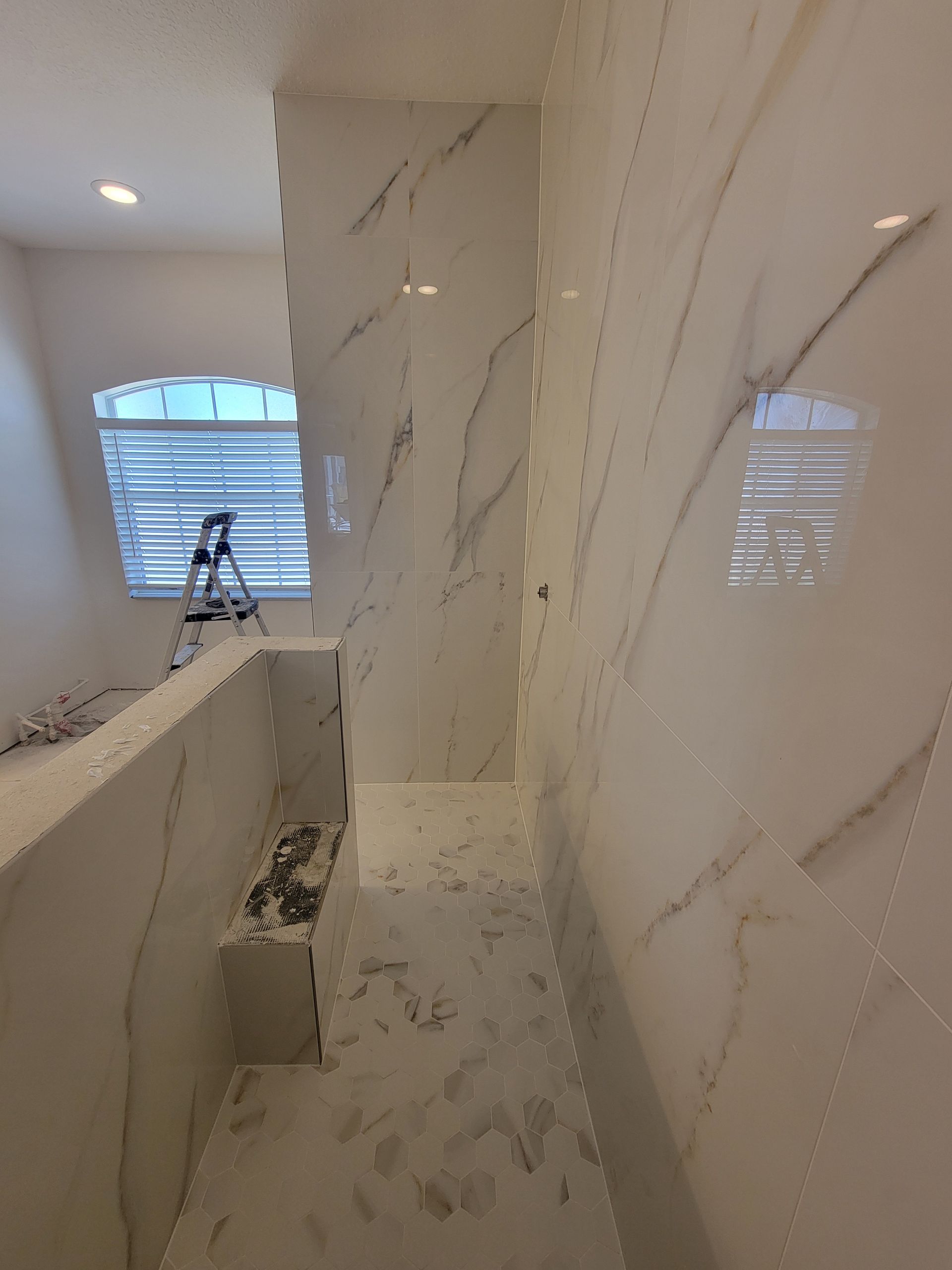 Bathroom with marble-like white tile. Window with blinds, a ladder, and unfinished steps are visible.